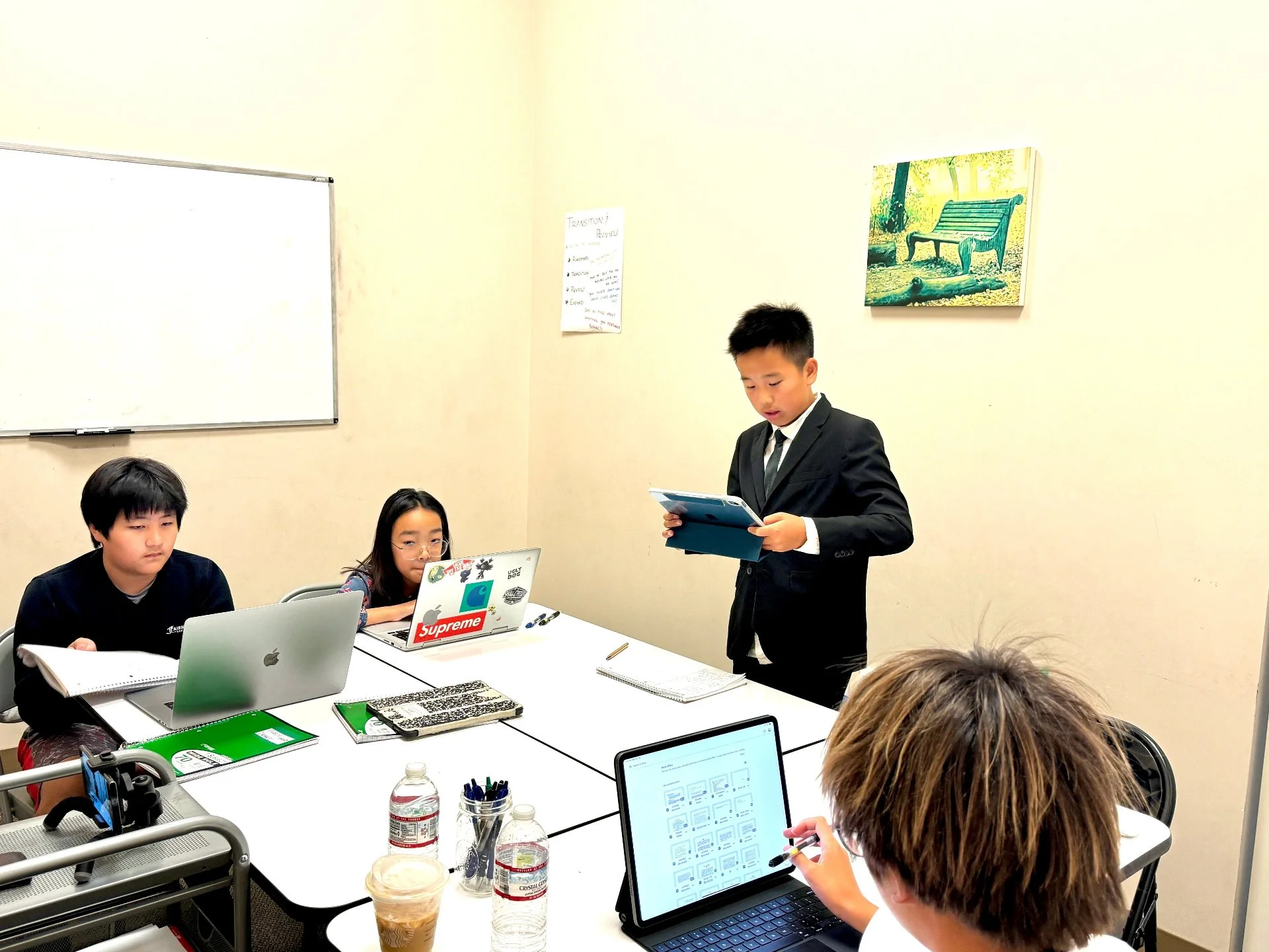 Students in a classroom with laptops and a student in a suit standing and speaking to the group.