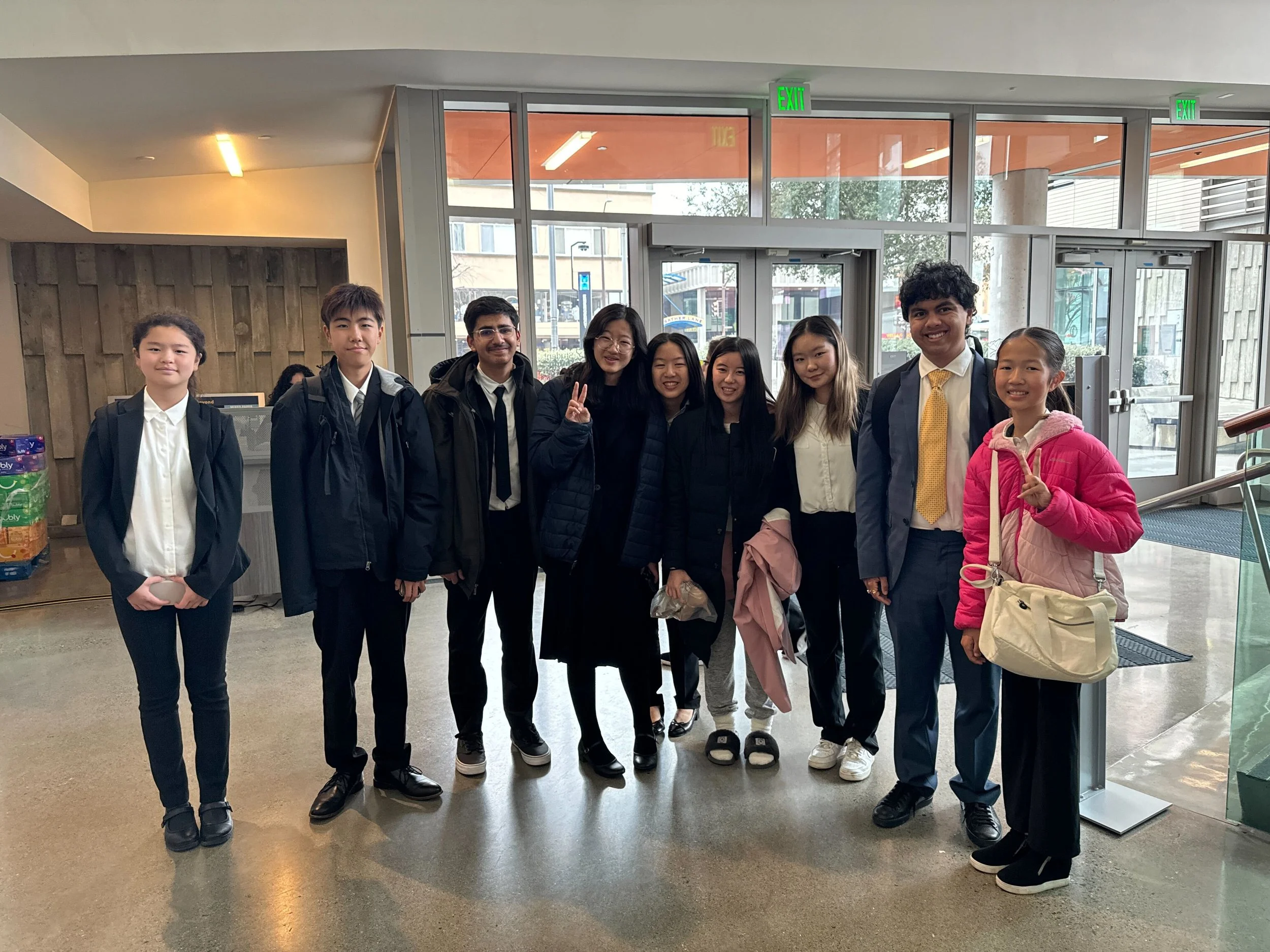 Group of nine students standing inside a building in front of glass doors, some smiling and making peace signs.