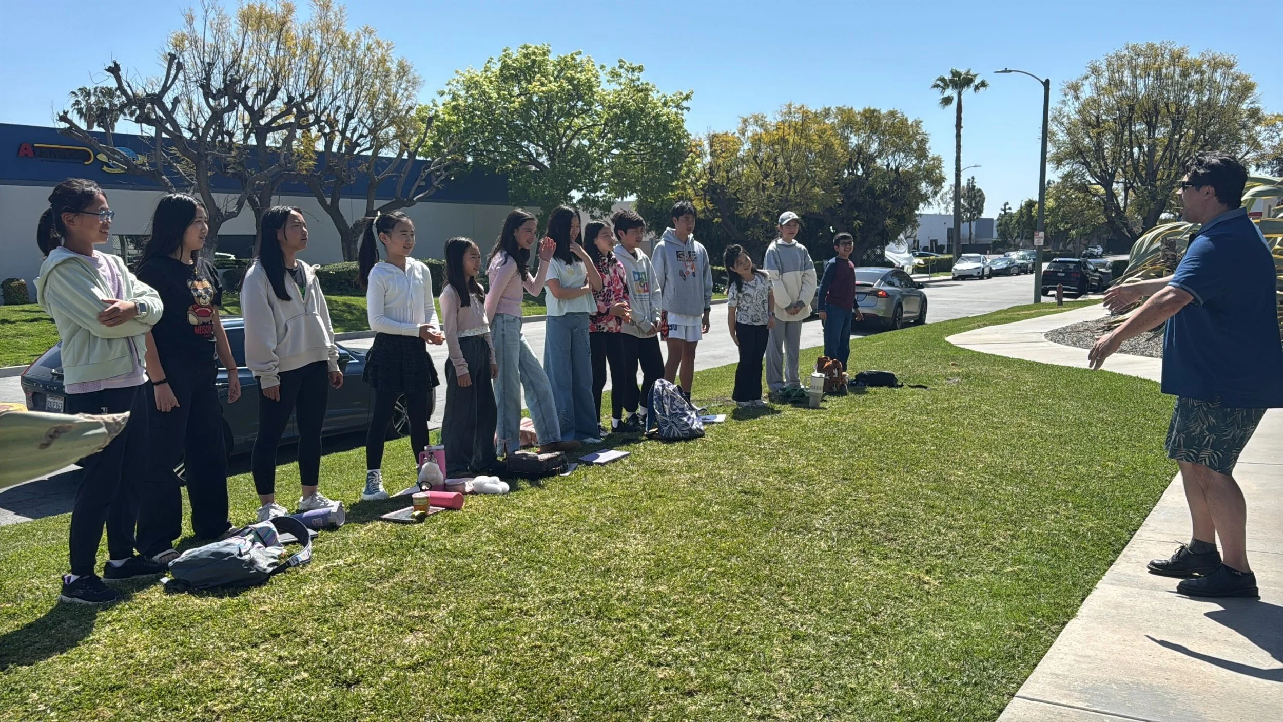 A group of young students standing on grass outdoors, facing an instructor who is talking to them. The students have backpacks and personal items on the ground in front of them. The weather is sunny with a clear blue sky, and there are trees, parked 