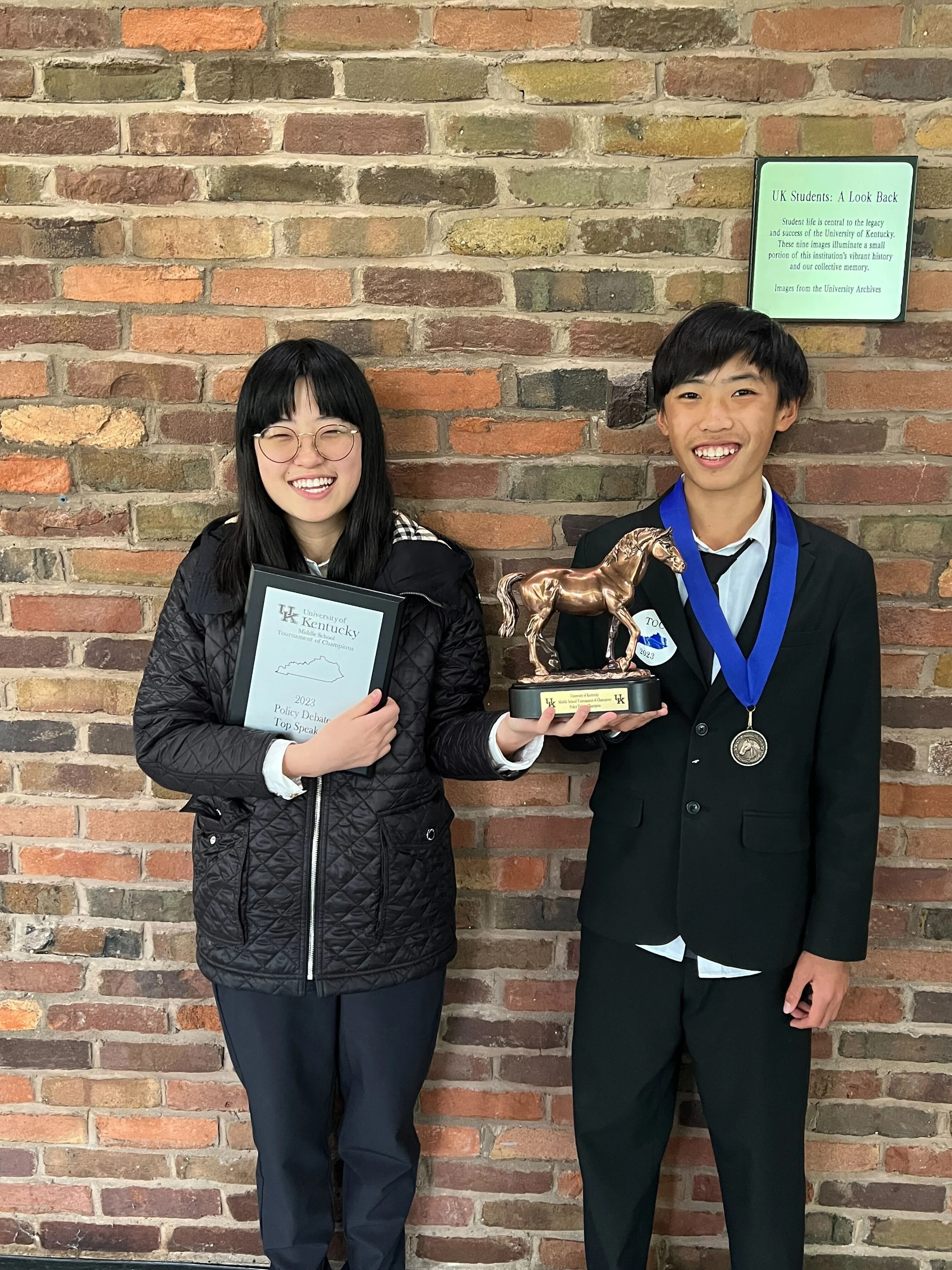 Two students standing in front of a brick wall, smiling and holding a trophy and a certificate. The student on the right is wearing a suit with a medal around their neck, and the student on the left is wearing glasses and a quilted jacket.