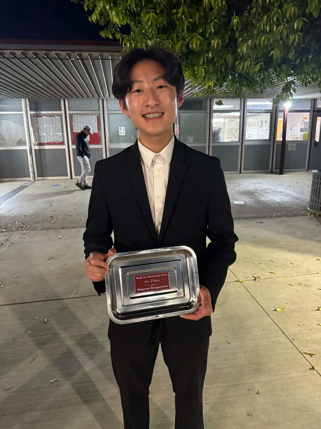 A young man in a black suit and white shirt smiling and holding a silver tray with a red award plaque that reads 'Woll Club International 2019 - 1st Place - Humorous Interpretation.' He is standing outside at night with a background of a building and