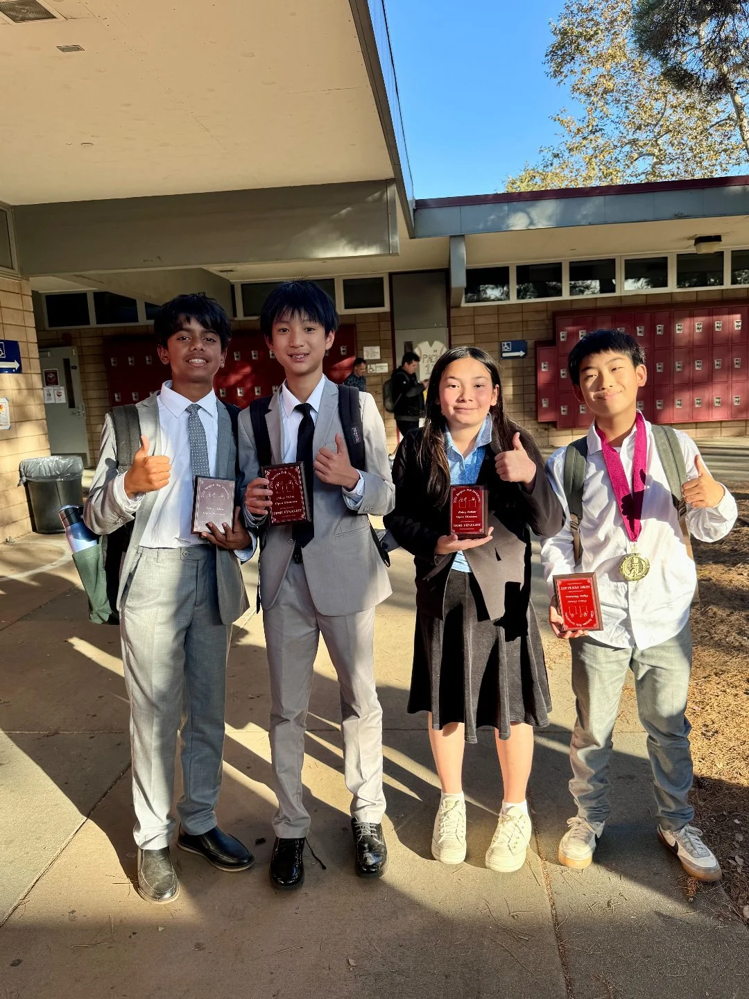 Four school students, two boys and two girls, standing outdoors in front of a school building, holding awards and giving thumbs up with a sunny sky above.