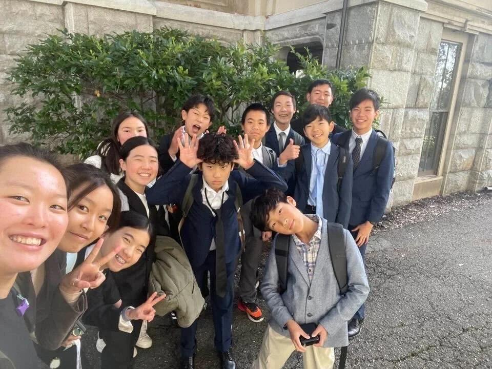 Group of school children and a teacher taking a selfie outside a stone school building, smiling and making peace signs.