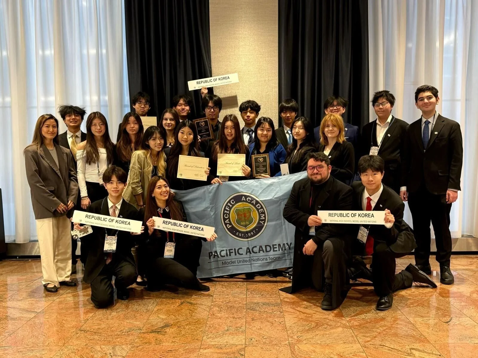 Group of young people in formal attire holding signs and certificates, posing behind a banner that reads 'Pacific Academy' and 'Model United Nations Team' in a conference room with curtains.