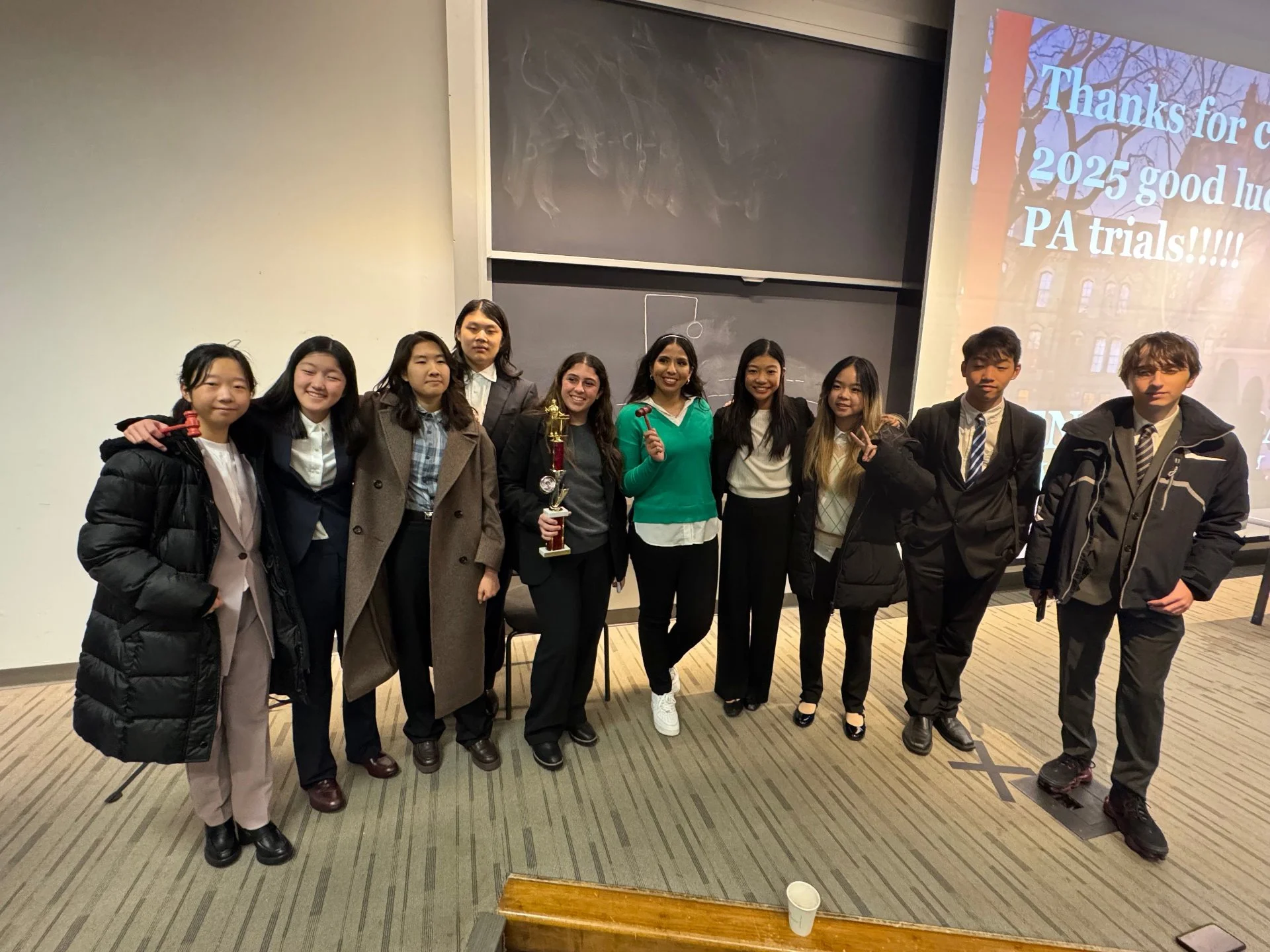 Group of ten young students standing together in a classroom, some holding a trophy, celebrating their achievement in Pennsylvania trials, with a presentation slide thanking for good luck in 2025 trials.