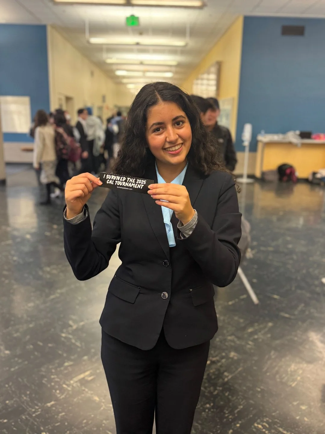 A young woman dressed in a black blazer holding a sticker that reads 'I SURVIVED THE 2025 CAL TOURNAMENT,' smiling in a school hallway with other students in the background.