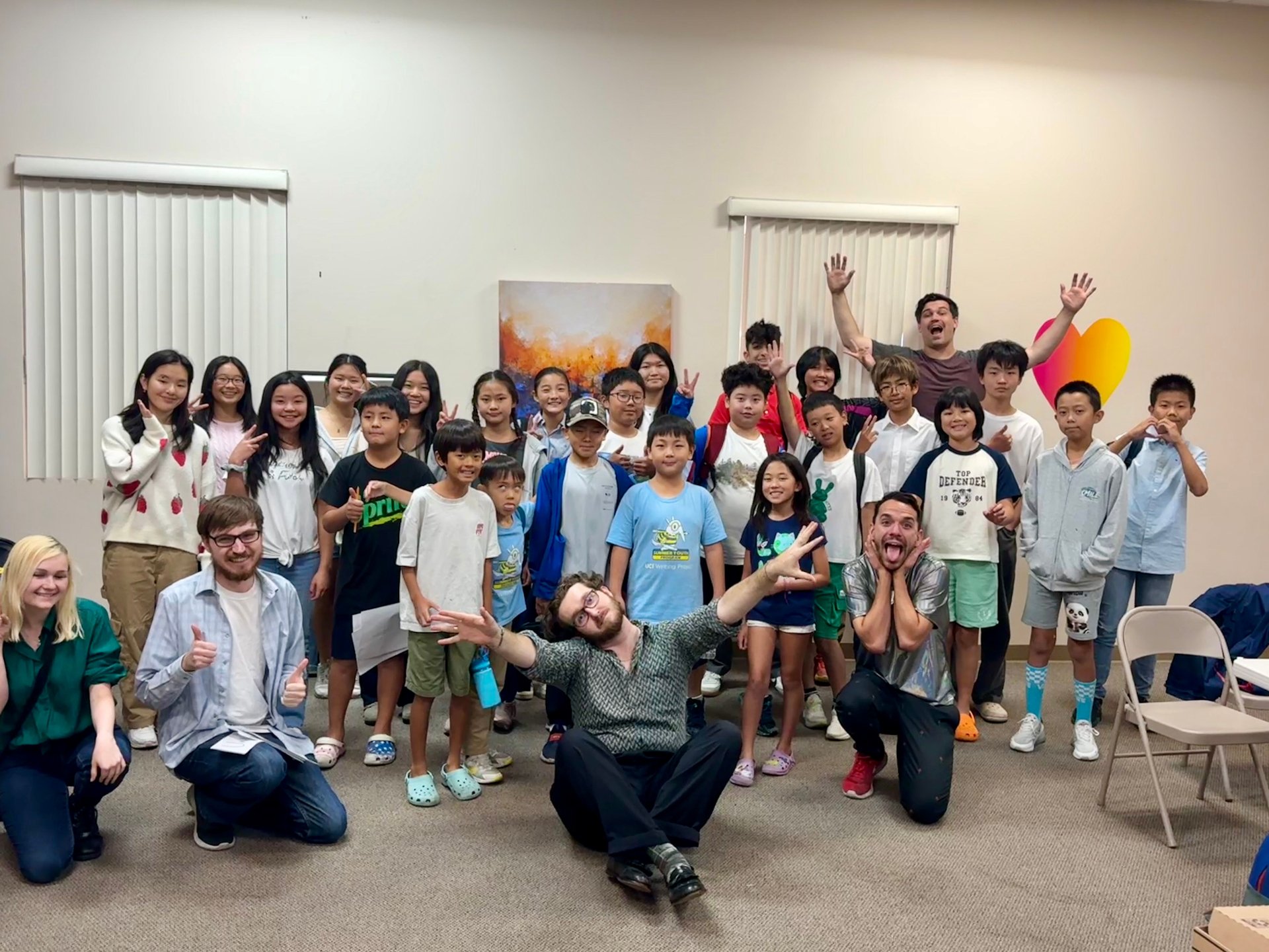 Group of children and young adults in a room posing for a group photo, some making peace signs and other playful gestures, with a man in the back raising his arms in excitement.
