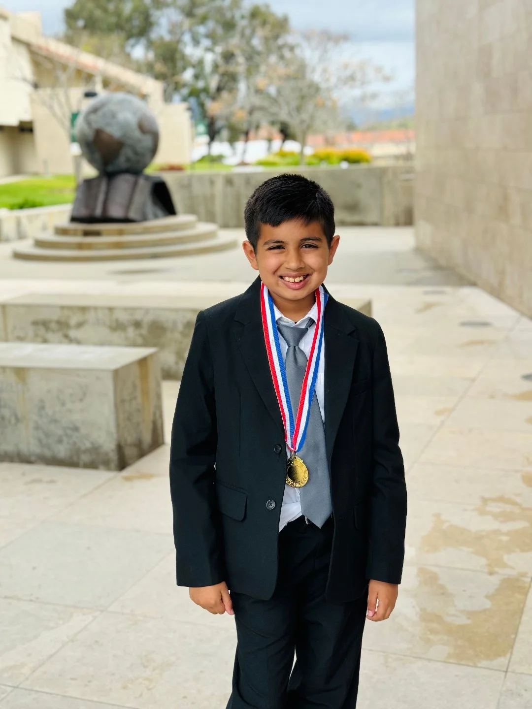 Young boy in a black suit, grey tie, and white shirt, wearing gold medal and Red, White, and Blue ribbon medals, smiling outdoors with a stone sculpture and trees in the background.