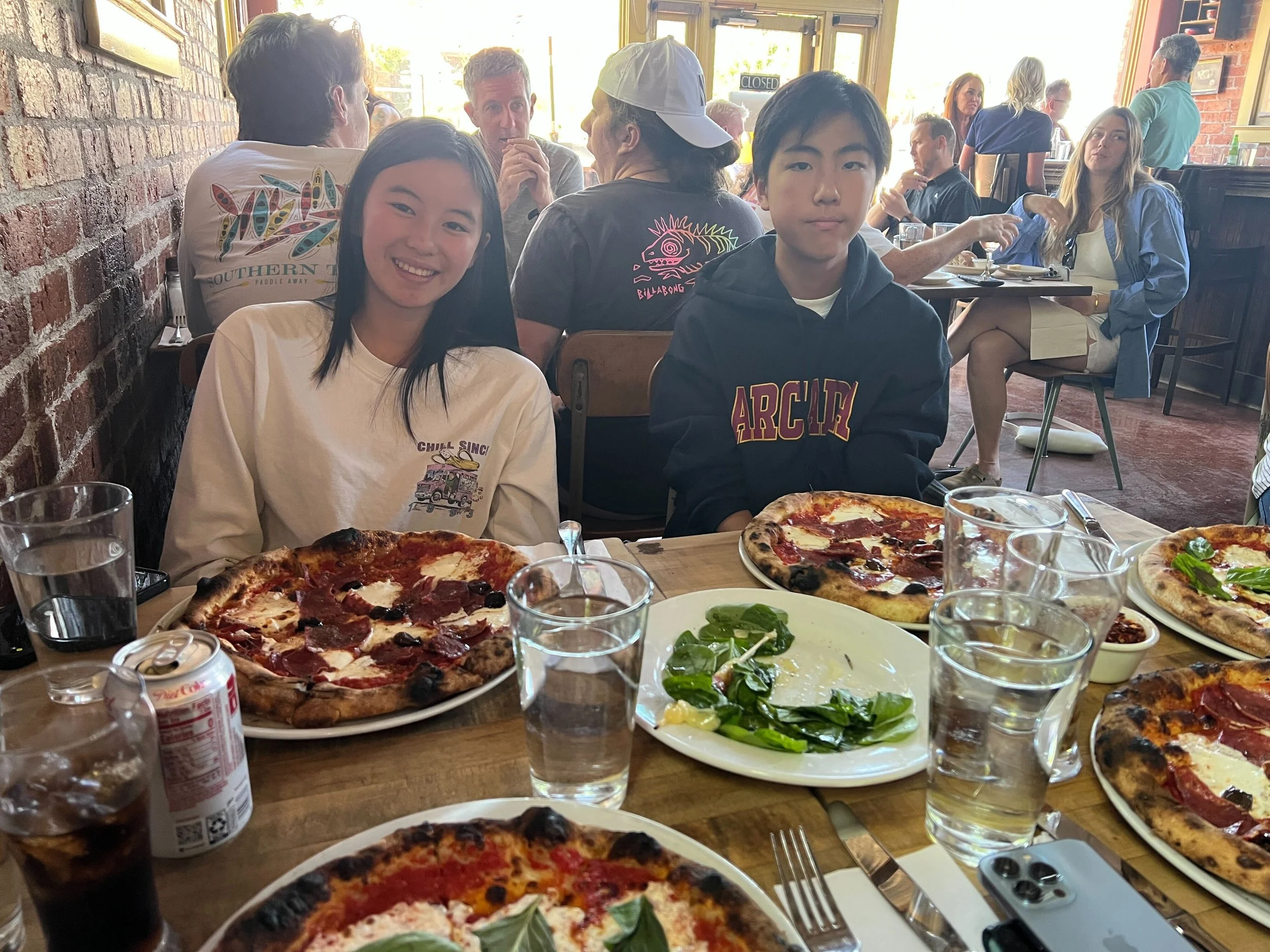 Two teenagers, a girl and a boy, sit at a table in a restaurant with pizza, salad, drinks, and a phone. The girl is smiling, and the boy has a neutral expression. There are other diners in the background.
