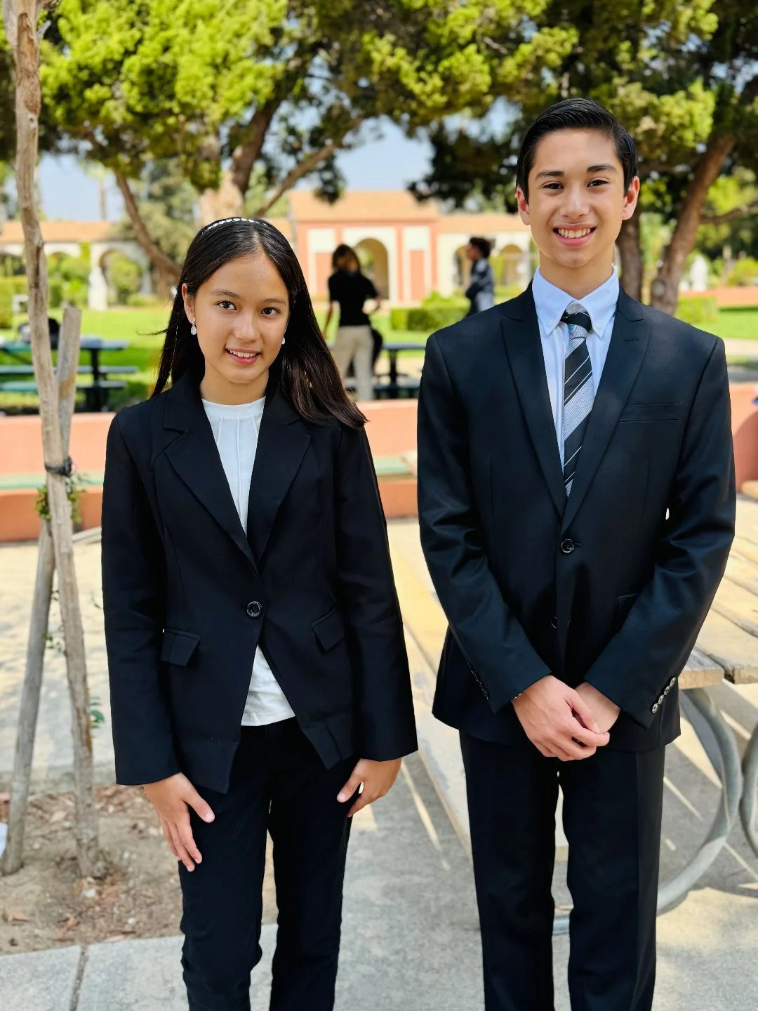 Two young people in formal suits standing outdoors in a park with trees and benches.