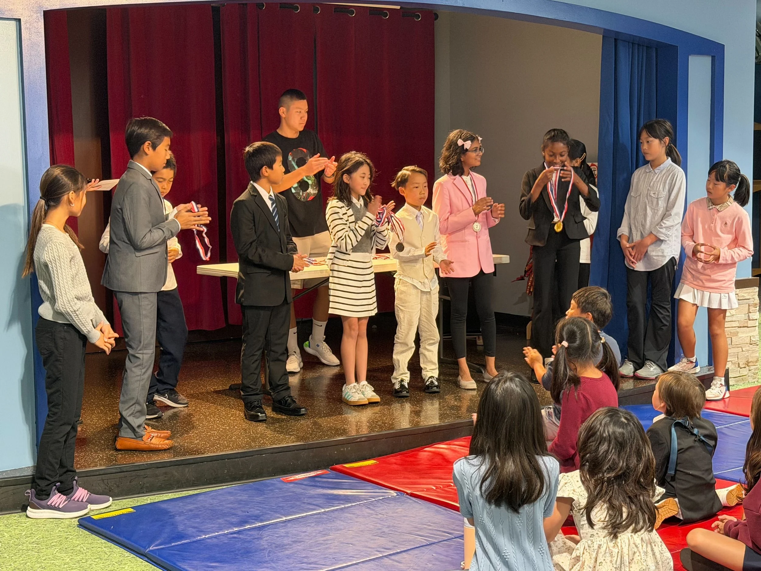 Children on a stage receiving medals and awards during a school awards ceremony, with seated children applauding in the foreground.