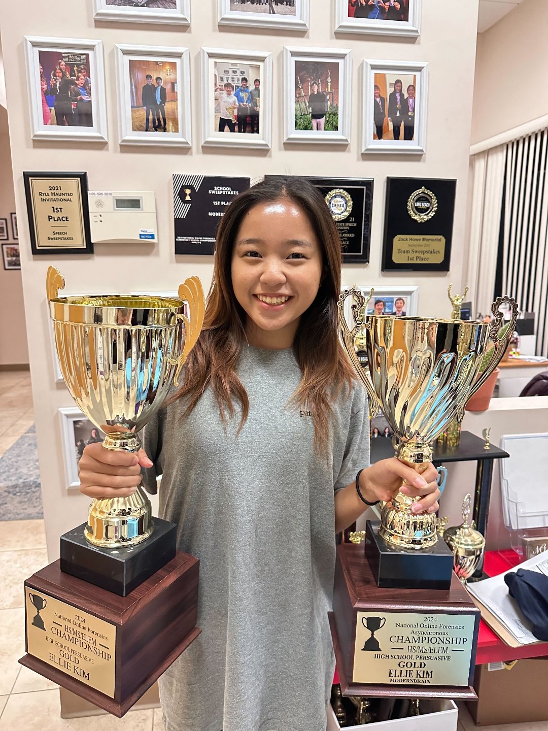 A young woman smiling and holding two large gold trophies, standing in front of a wall with framed awards and photographs.