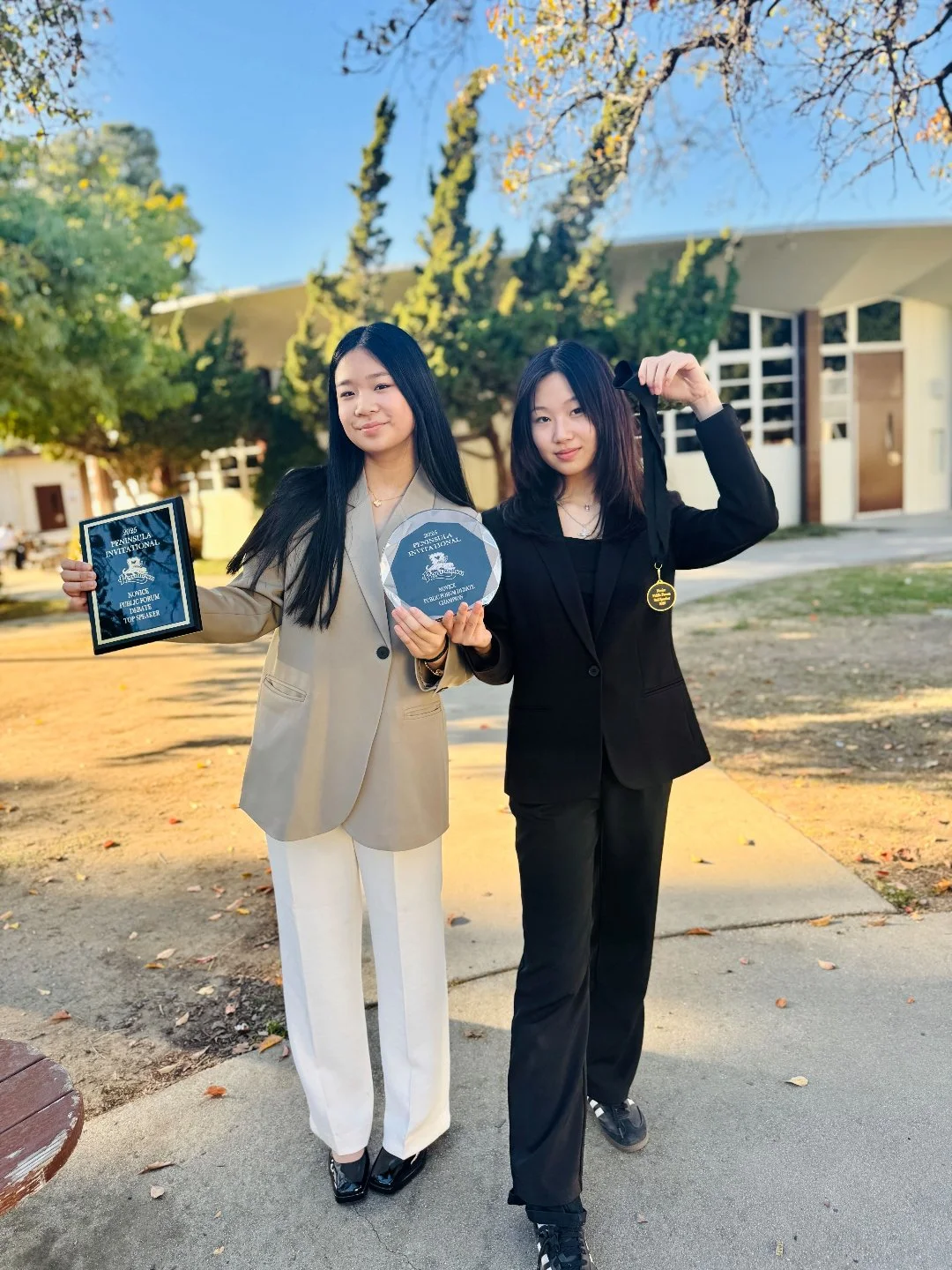 Two young women standing outdoors holding awards, one in a beige blazer and white pants, the other in a black blazer and pants, with trees and a modern building in the background.