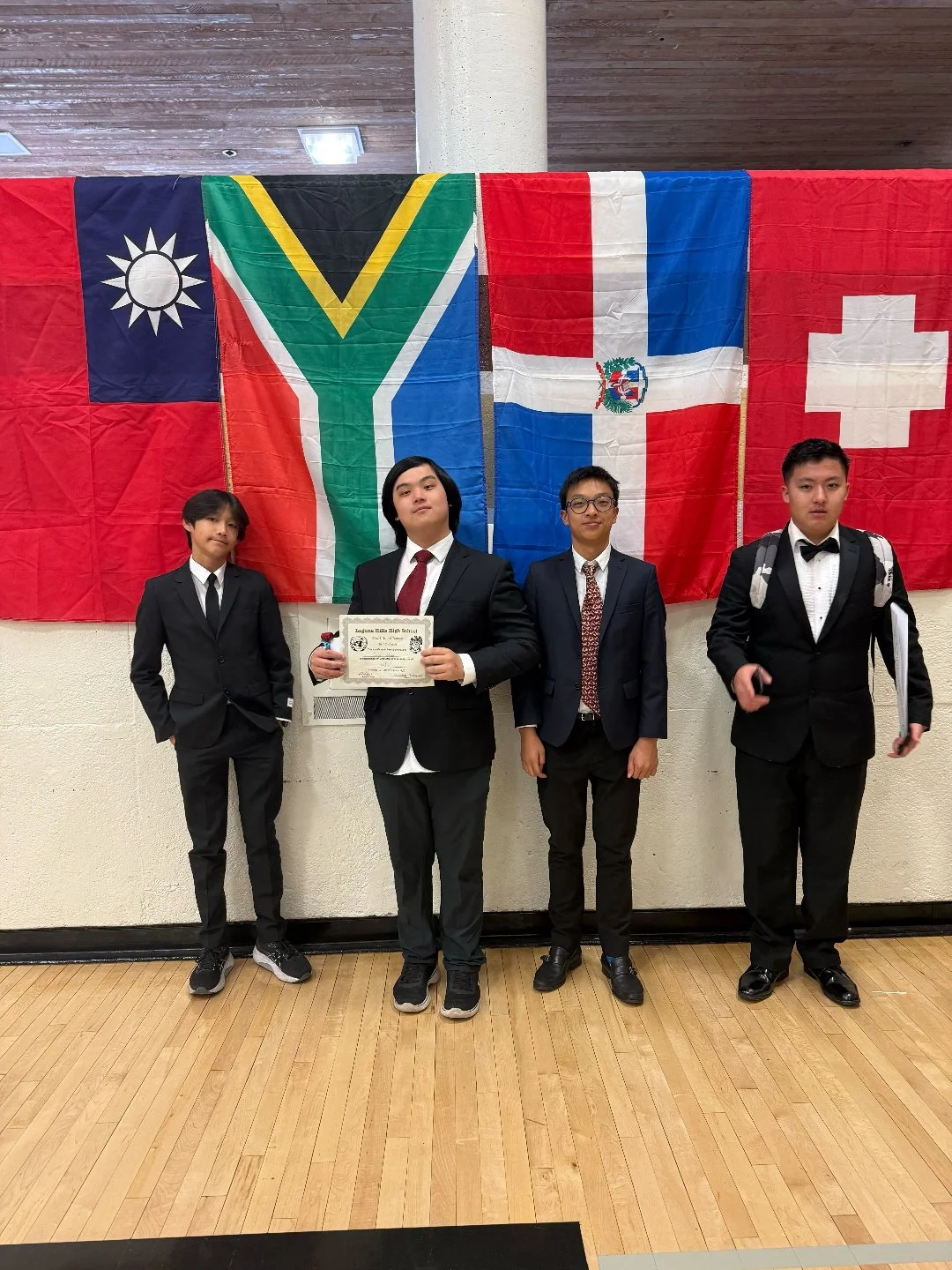 Four young men in formal suits standing in front of a display of four international flags, with one holding a certificate.