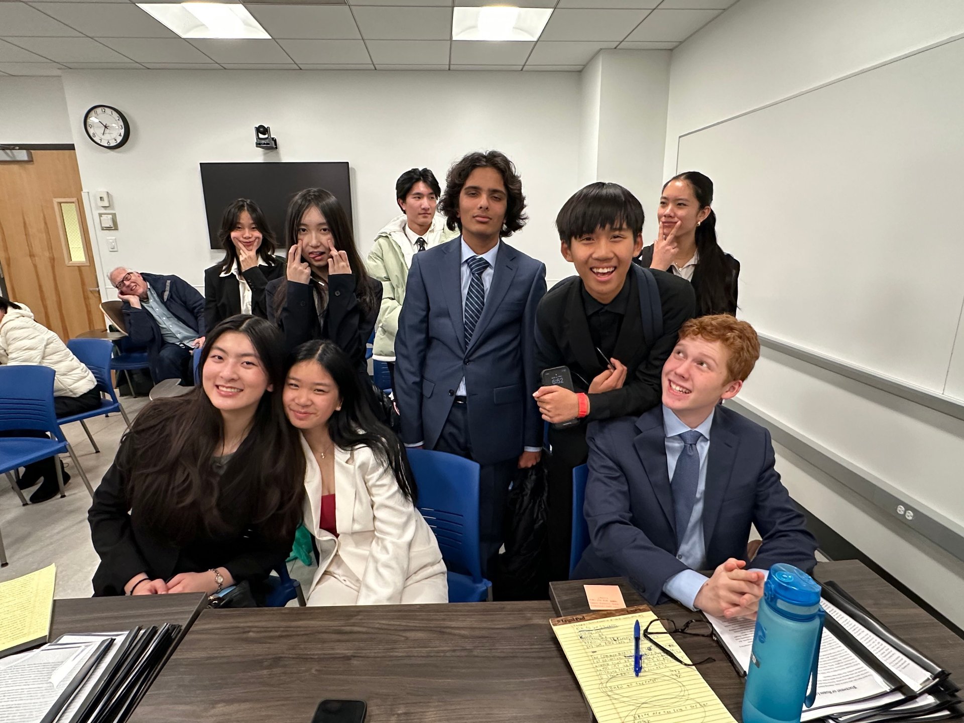 Group of young students and one adult in a classroom, some smiling at the camera, some making funny faces, with desks, notebooks, and water bottles visible.