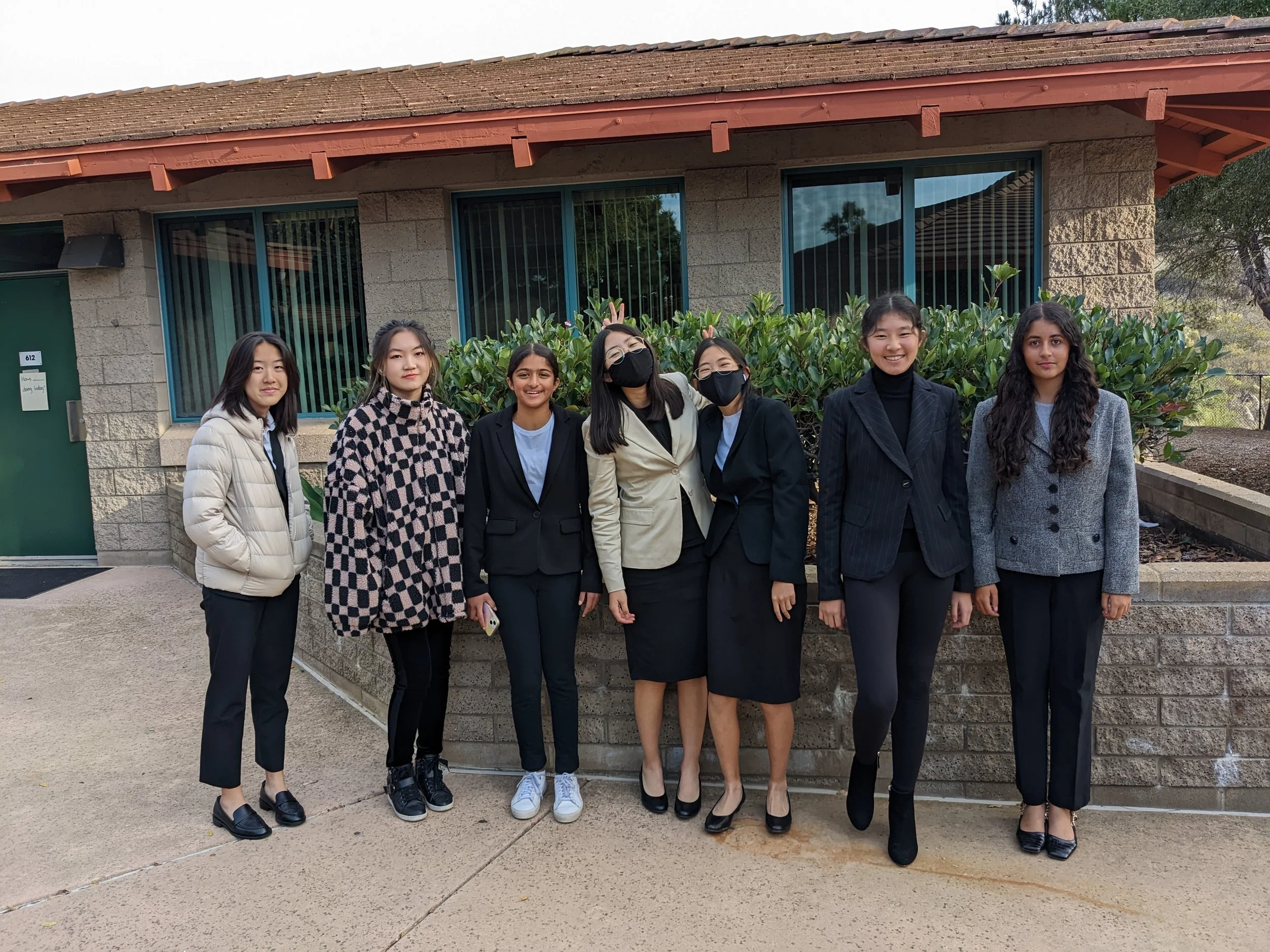 Group of seven young women standing outside in front of a building with bushes. They are dressed in semi-formal and casual clothing, some wearing masks.