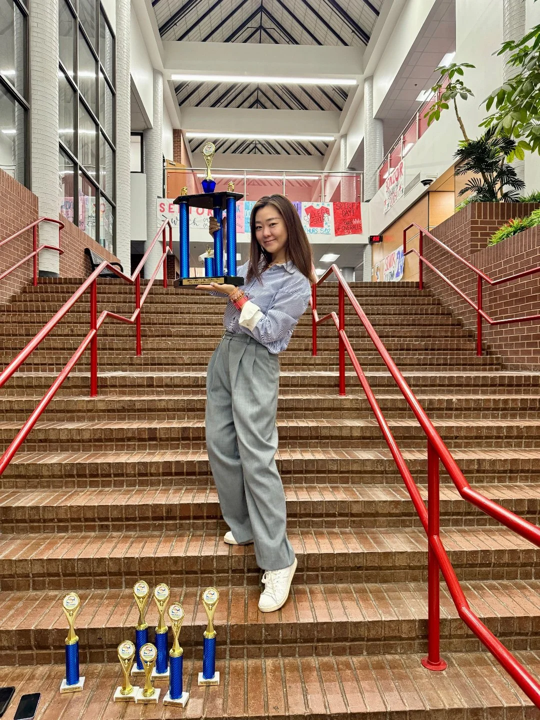 A young woman standing on a staircase indoors, holding a large blue and gold trophy, with several more trophies placed on the steps in front of her. She is smiling and dressed in a striped shirt and gray high-waisted pants.