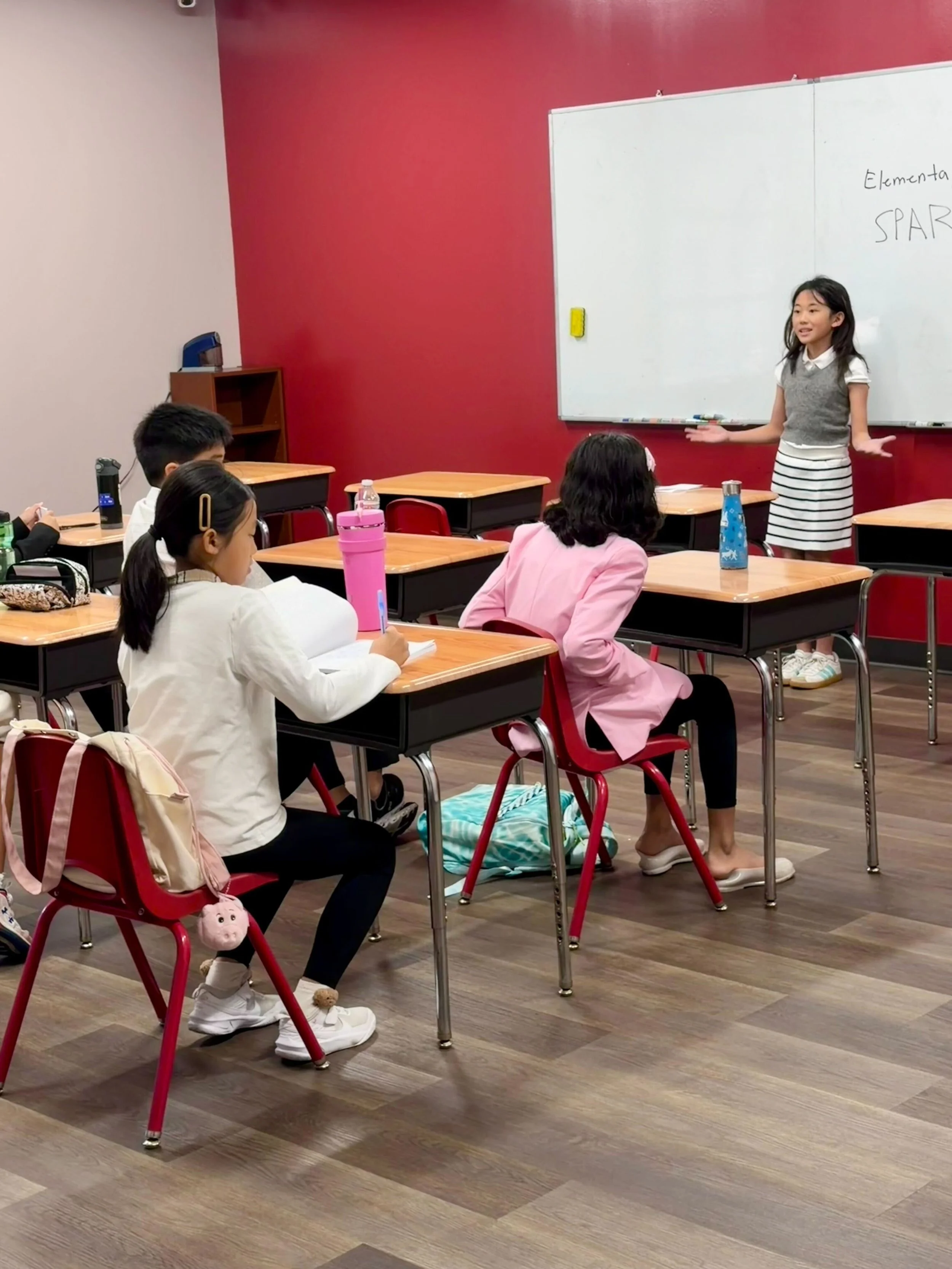 A classroom with five young students listening to a girl presenting at the whiteboard, with a red wall background, and desks with water bottles and backpacks.