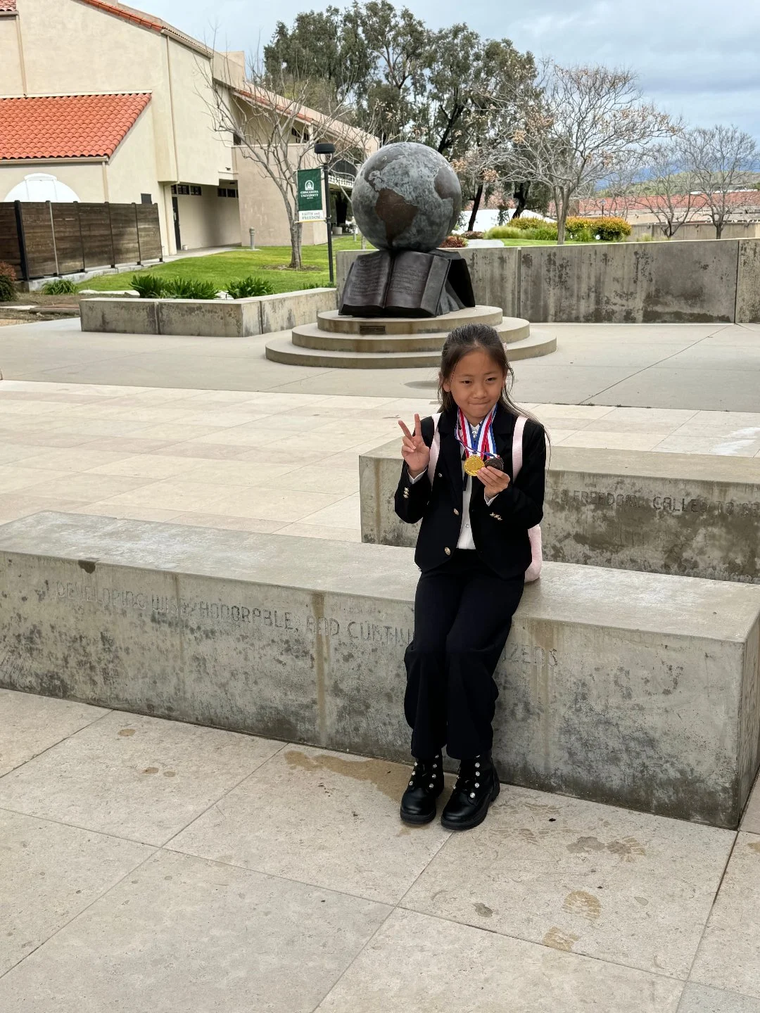 A young girl in a black school uniform with medals around her neck, sitting on a concrete bench at a school campus, making a peace sign, with a globe and open book sculpture in the background.