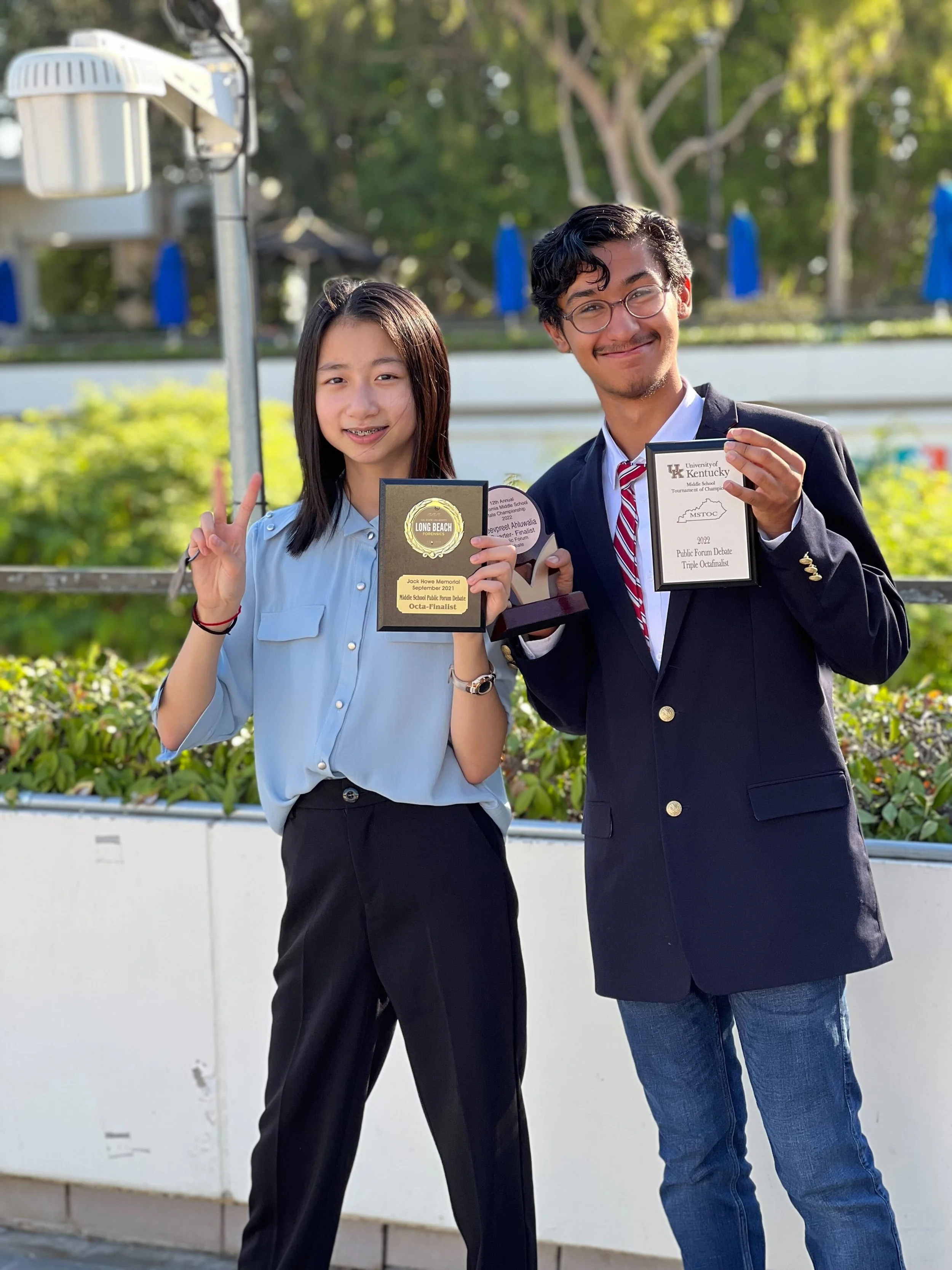 Two young students, a girl and a boy, standing outdoors, smiling, holding awards and making peace signs. The girl is wearing a light blue blouse and dark pants, and the boy is in a dark blazer with a red striped tie and jeans. They are celebrating th