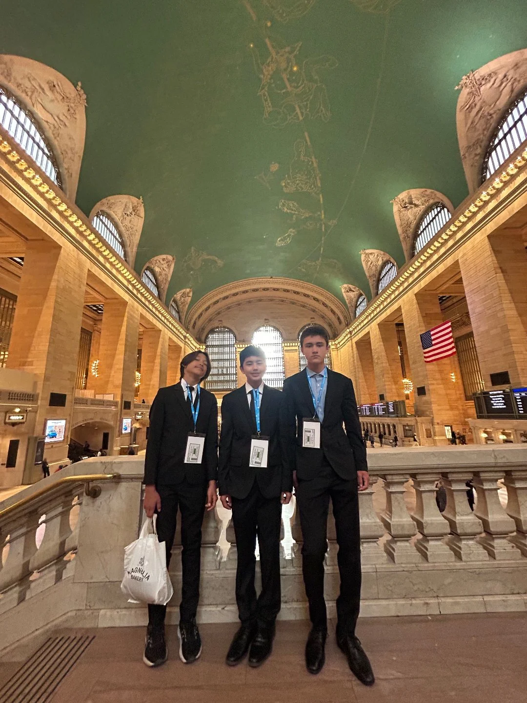 Three young men in formal suits standing on the stairs inside a grand, historic train station with high arched windows, a green decorated ceiling, and an American flag. They are wearing conference badges around their necks, indicating they are attend