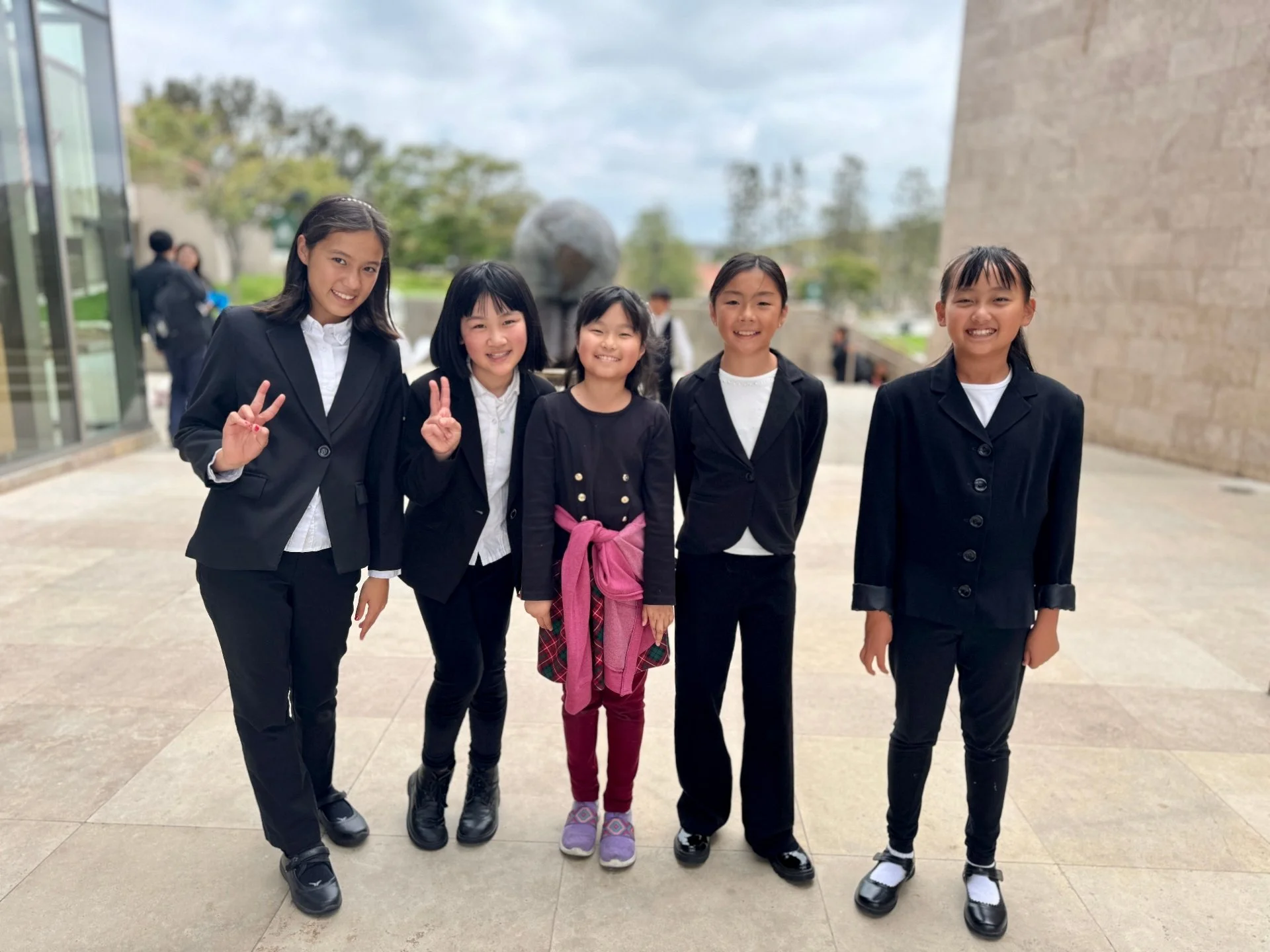 Five young girls dressed in black suits, standing outdoors and smiling at the camera.
