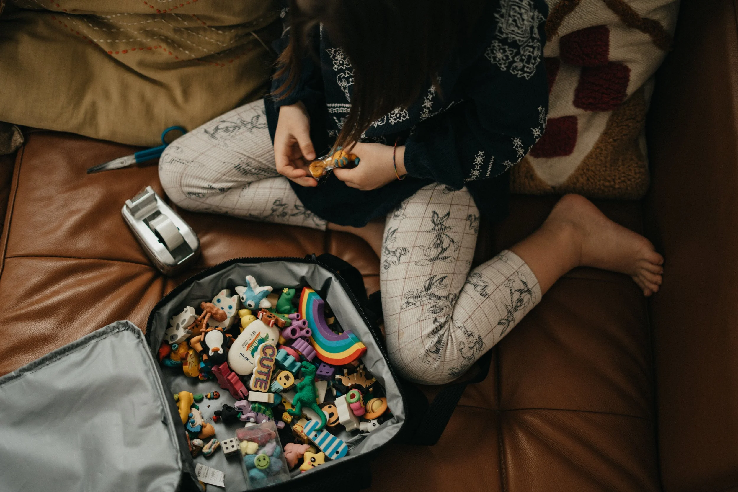 Ein Mädchen sitzt auf einer Ledercouch und spielt mit buntem Spielzeug, das in einem offenen Rucksack liegt.