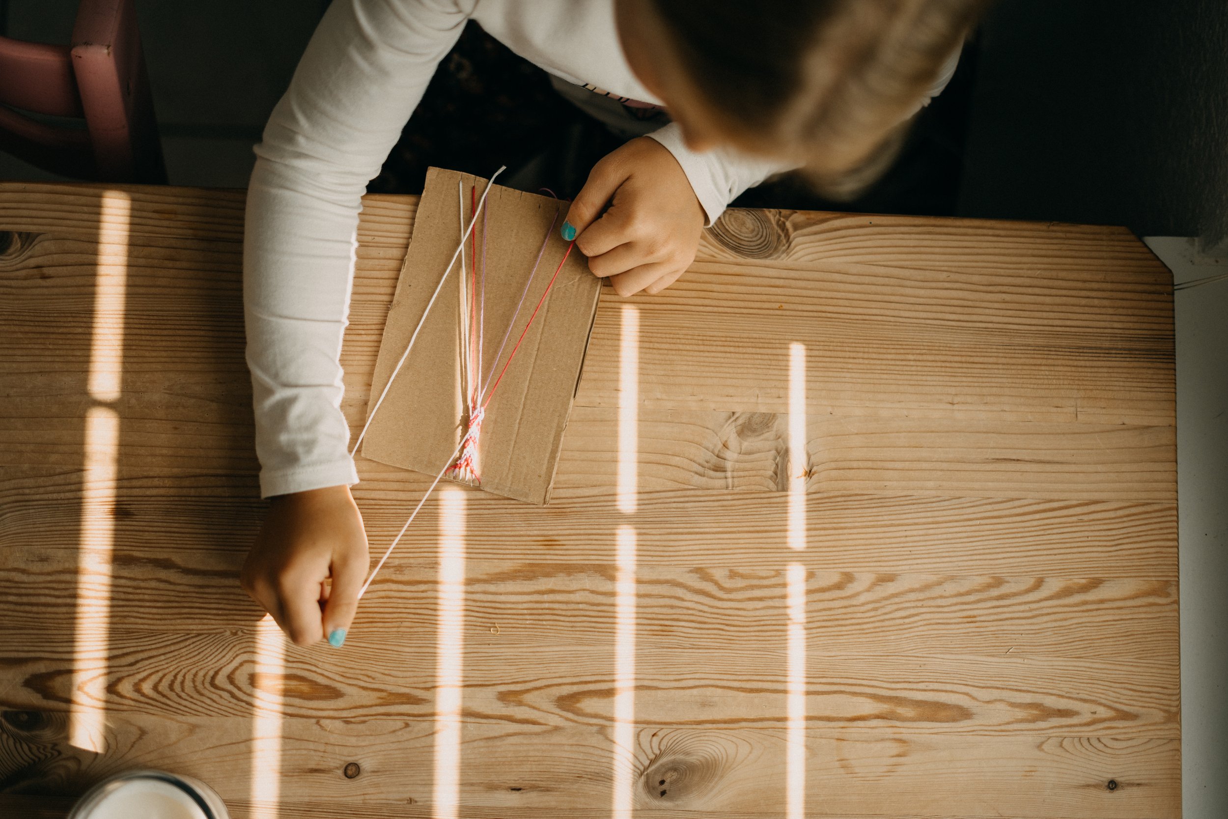 Ein Kind spielt mit einem Bastelset auf einem Holztisch, das bunte Fäden enthält.