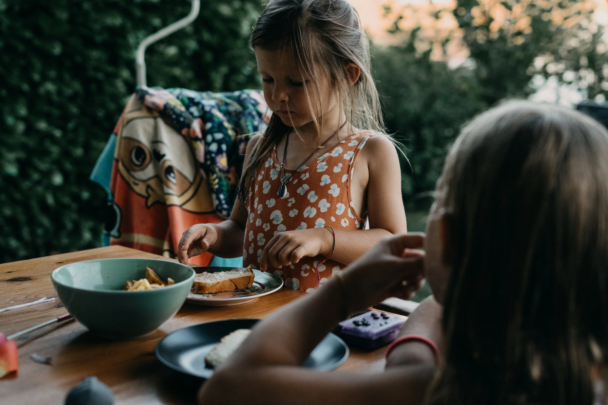 Authentische Dokumentarfotografie von zwei Kindern, die im Sommer draussen am Gartentisch sitzen und eine Zwischenmahlzeit geniessen.