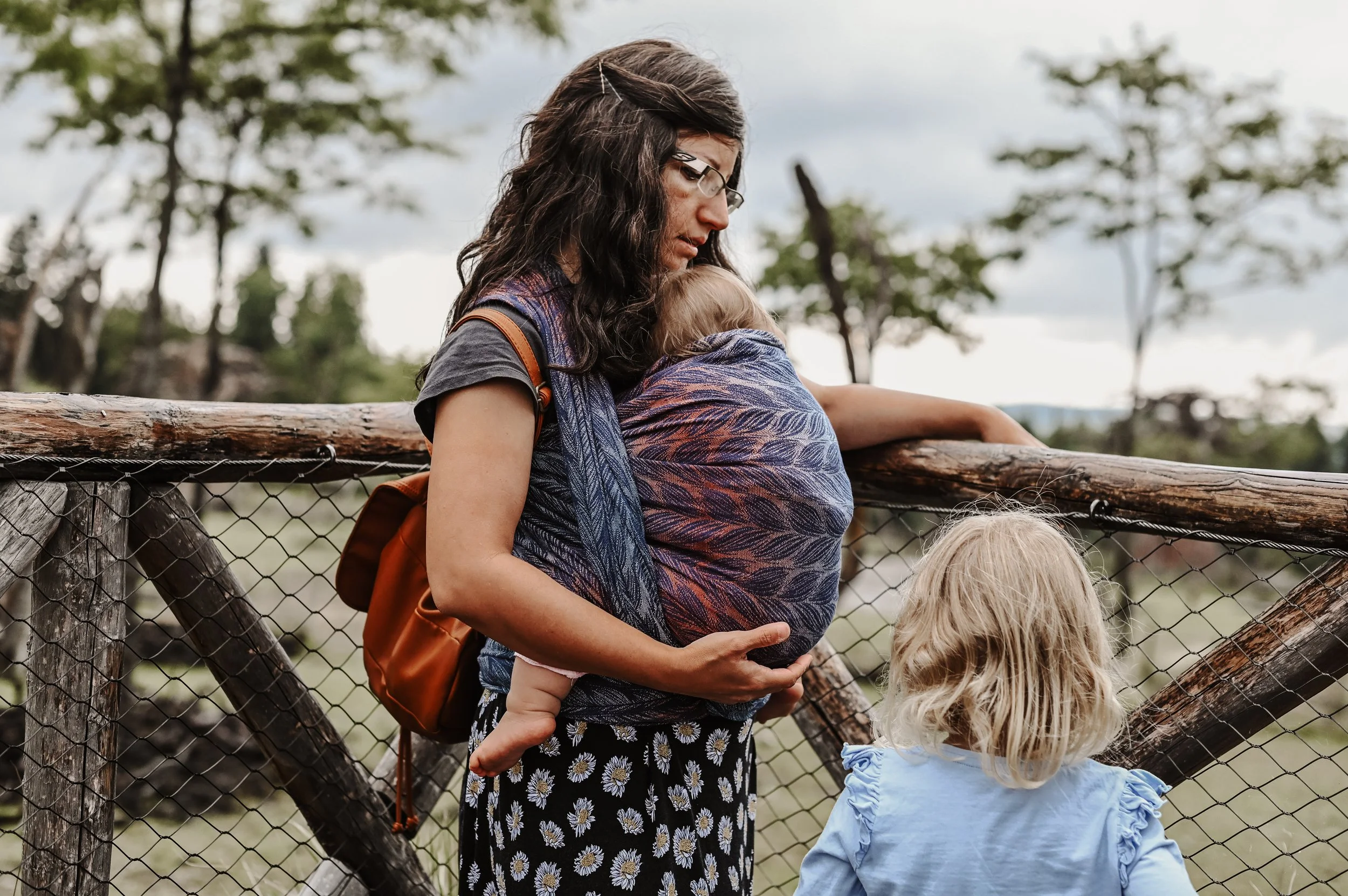 Eine Frau mit dunklen, gewellten Haaren trägt ein Baby in einer Trage und hält sich an einem Holzzaun fest, während ein Mädchen mit blonden, lockigen Haaren zu ihr schaut.