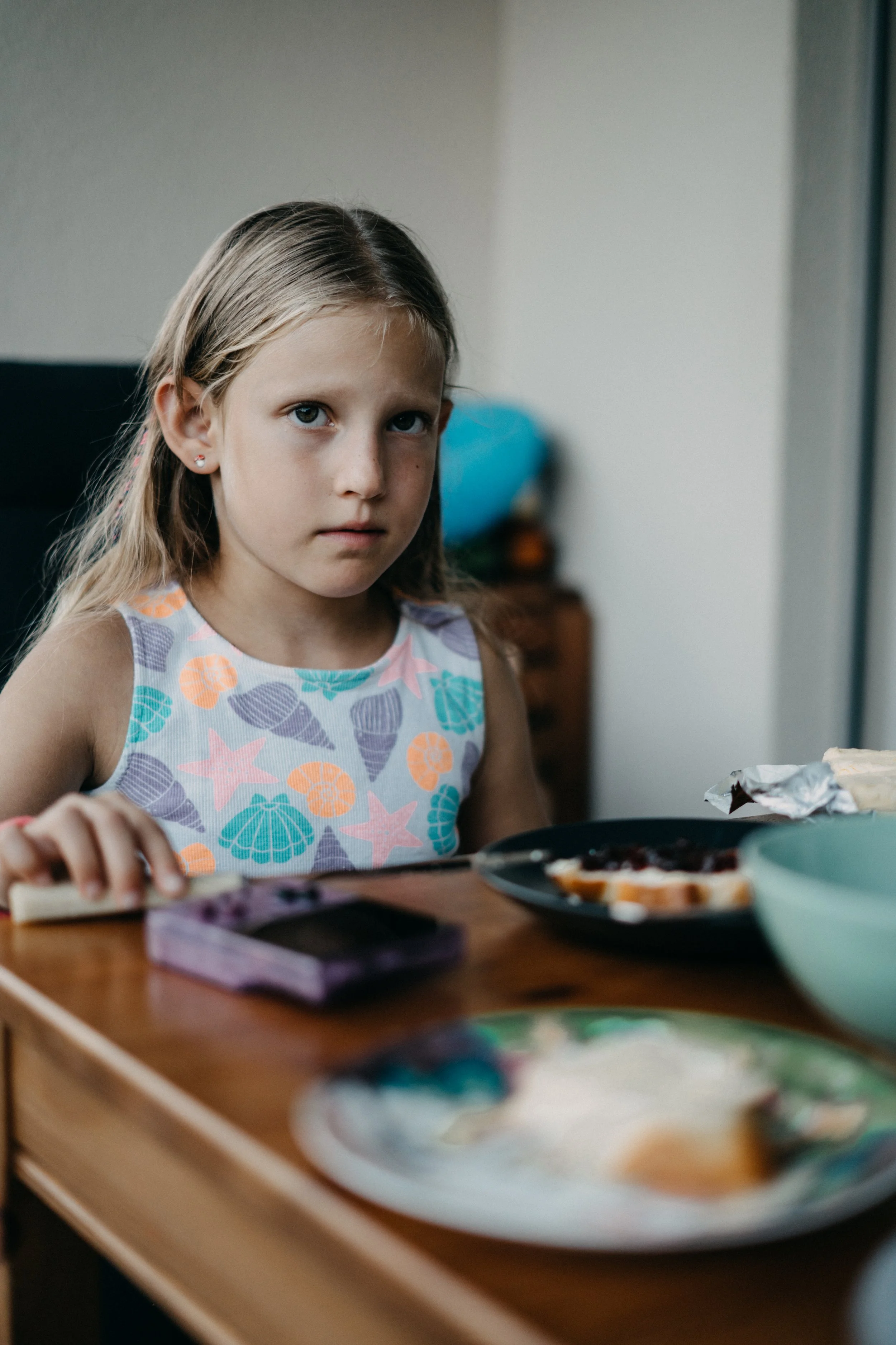 Ein Mädchen mit langem, blondem Haar, trägt ein ärmelloses Oberteil mit Muschel- und Sternenmustern, sitzt an einem Esstisch mit Tellern und Lebensmitteln, schaut ernst in die Kamera.