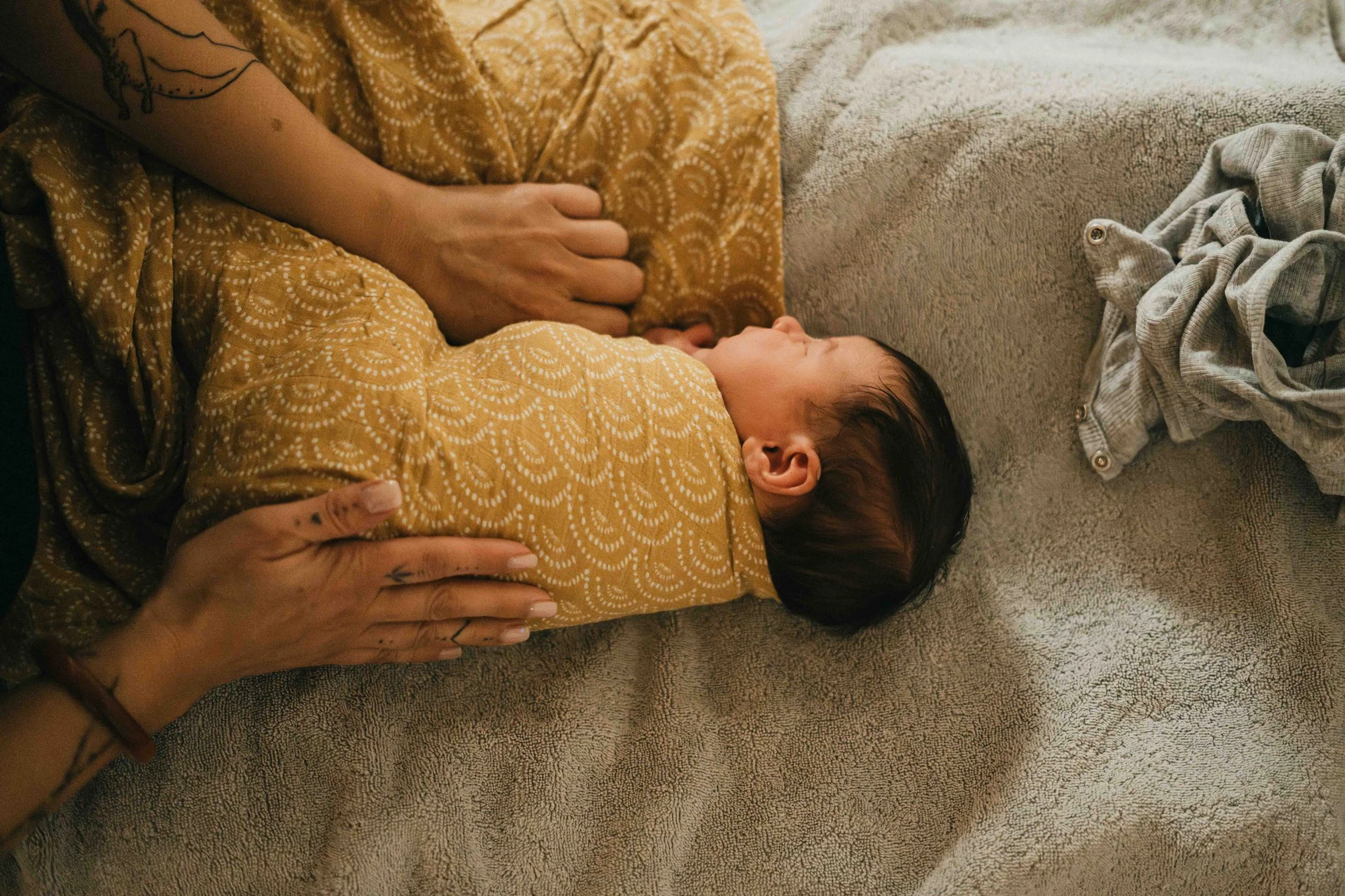 Ein Baby liegt auf einem weichen, hellen Handtuch auf dem Bauch, während eine Person mit Tattoos eine Hand auf den Rücken des Babys legt. Das Baby trägt eine gelbe, gemusterte Kleidung und ist momentan ruhig.