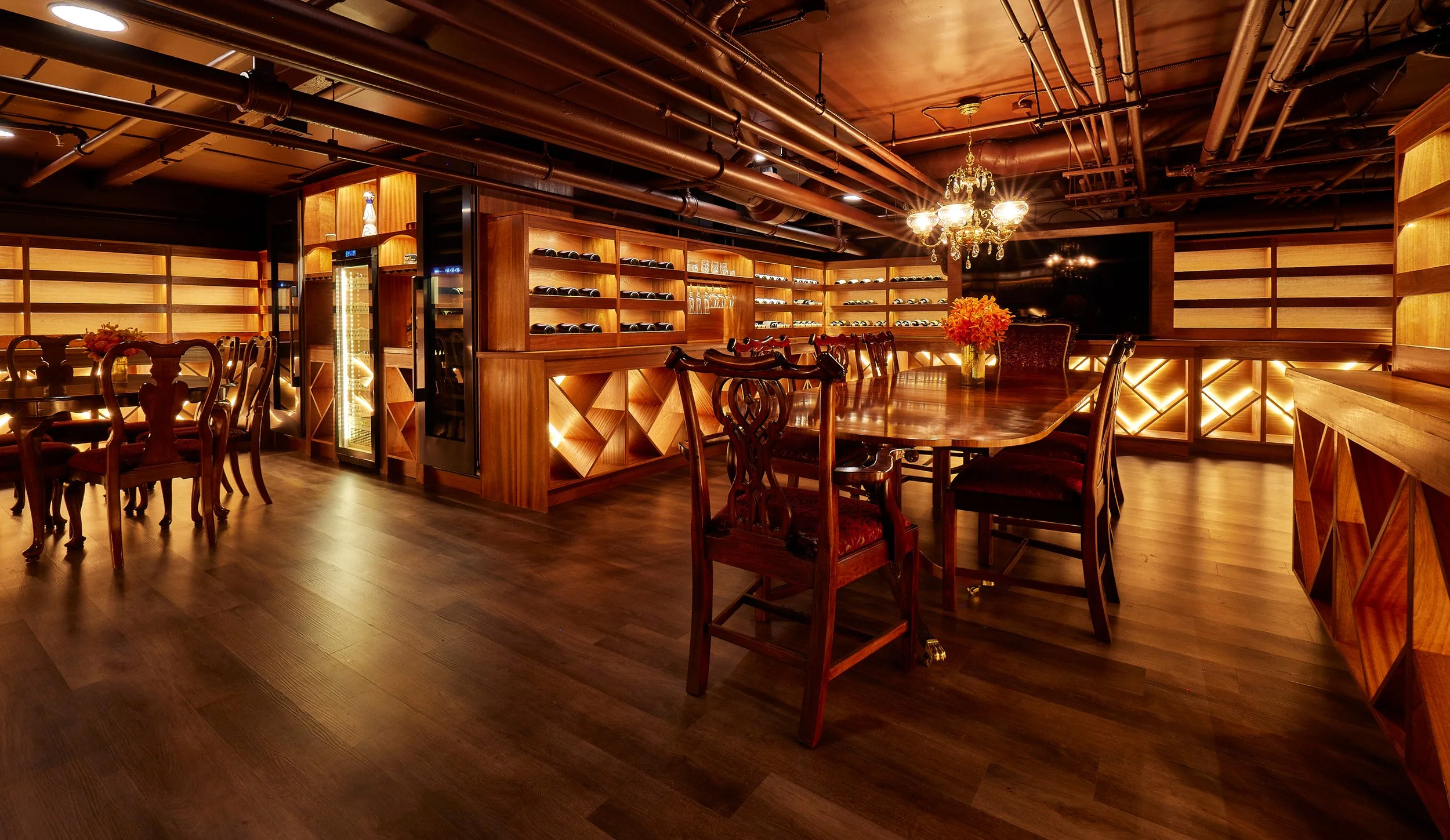 An elegant restaurant with wooden furniture, a chandelier, and wine bottles on shelves. The room has dim lighting and decorative flowers on the tables.
