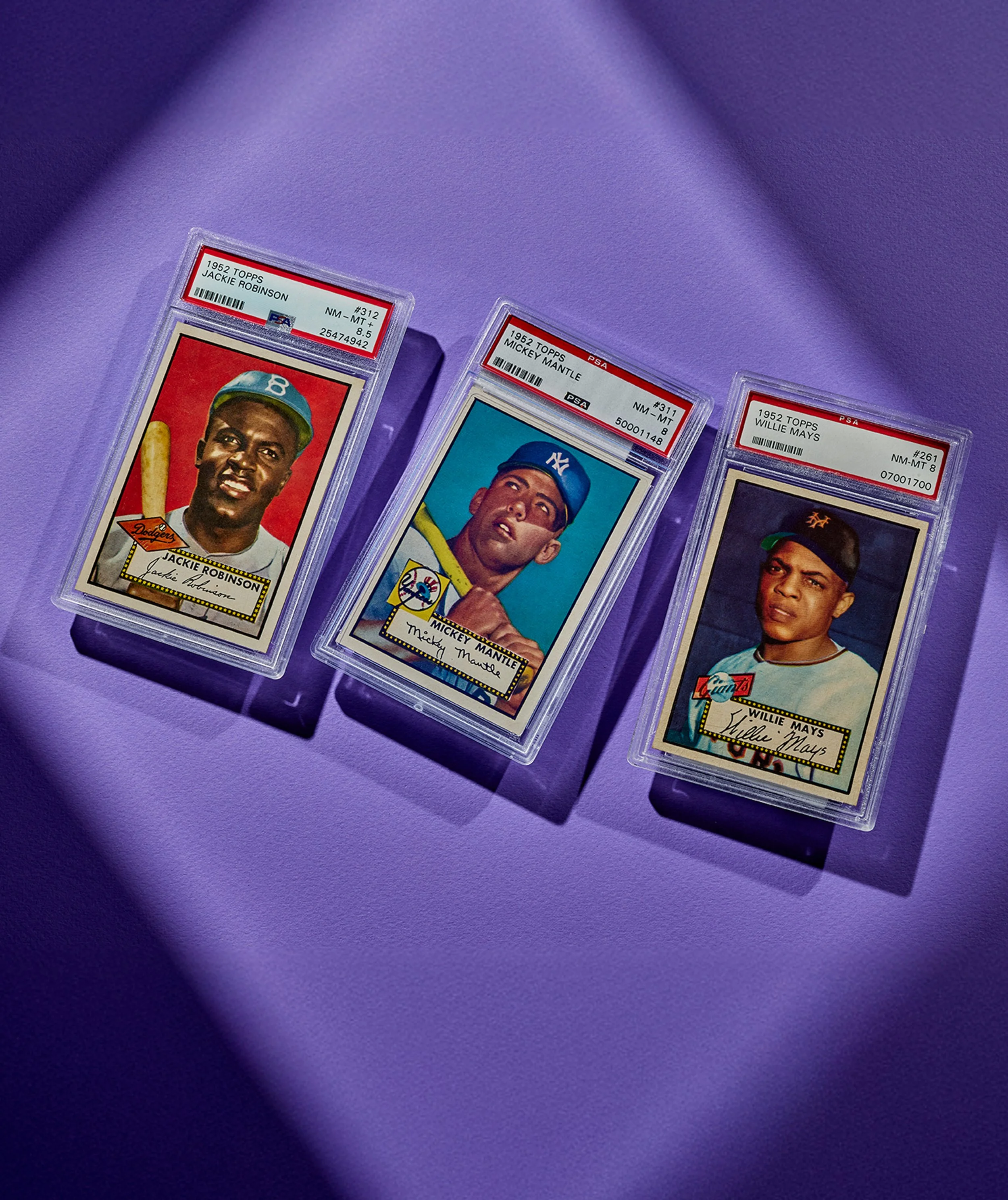 Three vintage baseball trading cards from 1952 featuring Jackie Robinson, Mickey Mantle, and Willie Mays, each in protective plastic cases, placed on a purple surface.