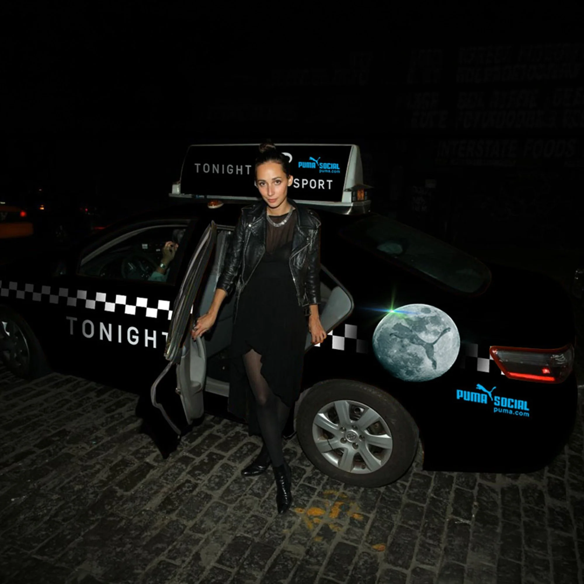 A woman with dark hair in a bun, wearing a leather jacket, standing next to a black-and-white checkered taxi car at night. The taxi has a sign on top reading 'TONIGHT' and 'SPORT' with a blue 'Puma Social' logo. The woman is holding onto the open car door, and there is a moon graphic at the bottom right corner of the image.