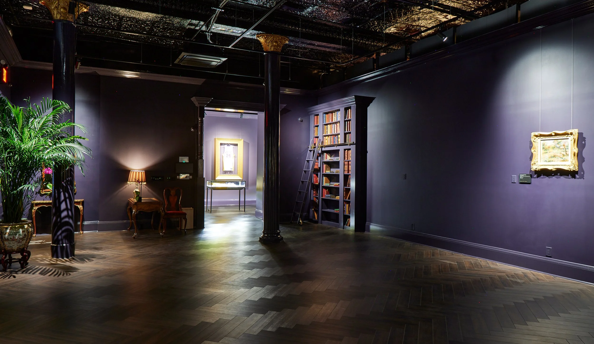 An elegant library or gallery room with dark purple walls, a ceiling with a reflective metallic surface, and polished wooden parquet flooring. There are tall black columns supporting the ceiling, a tall green houseplant on the left, and a small desk with a lamp and a red chair in the background. A bookshelf filled with books and a ladder are on the right side of the image, with some framed artwork hanging on the walls.