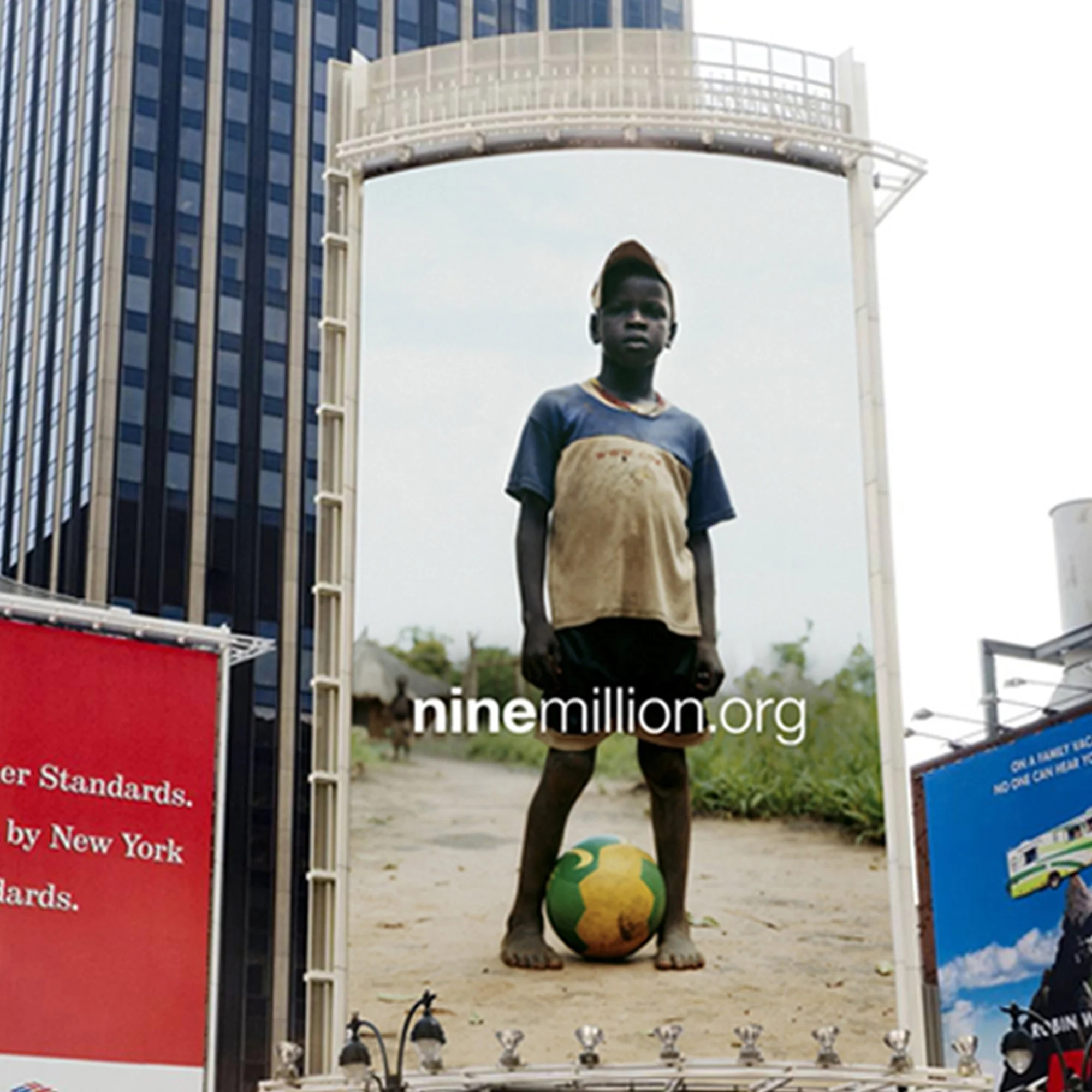 A young boy standing on a sidewalk, holding a green and yellow soccer ball, with a large urban billboard behind him in a city setting.
