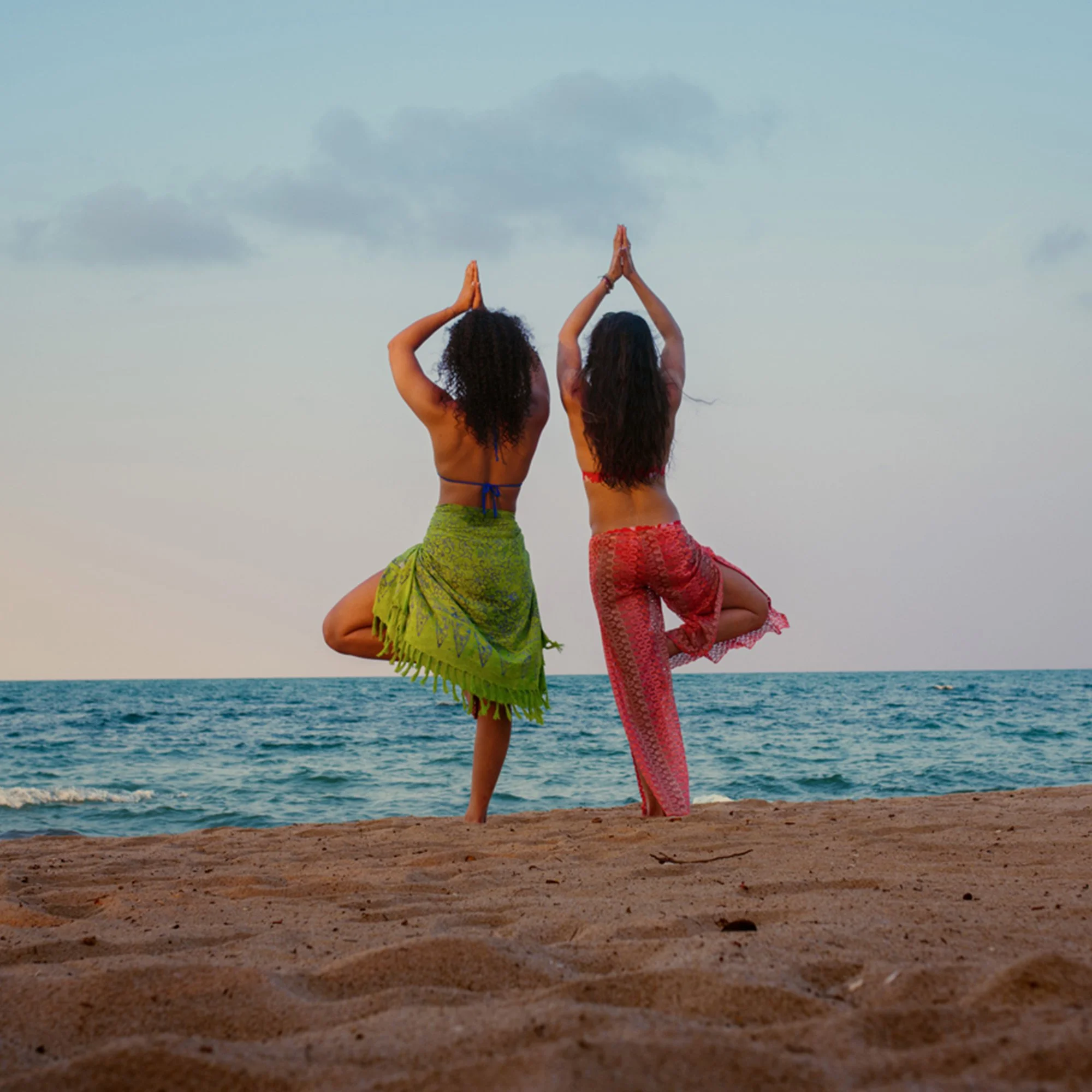 Two women practicing yoga on the beach near the ocean, standing on one leg with hands in prayer position above their heads, facing away from the camera.