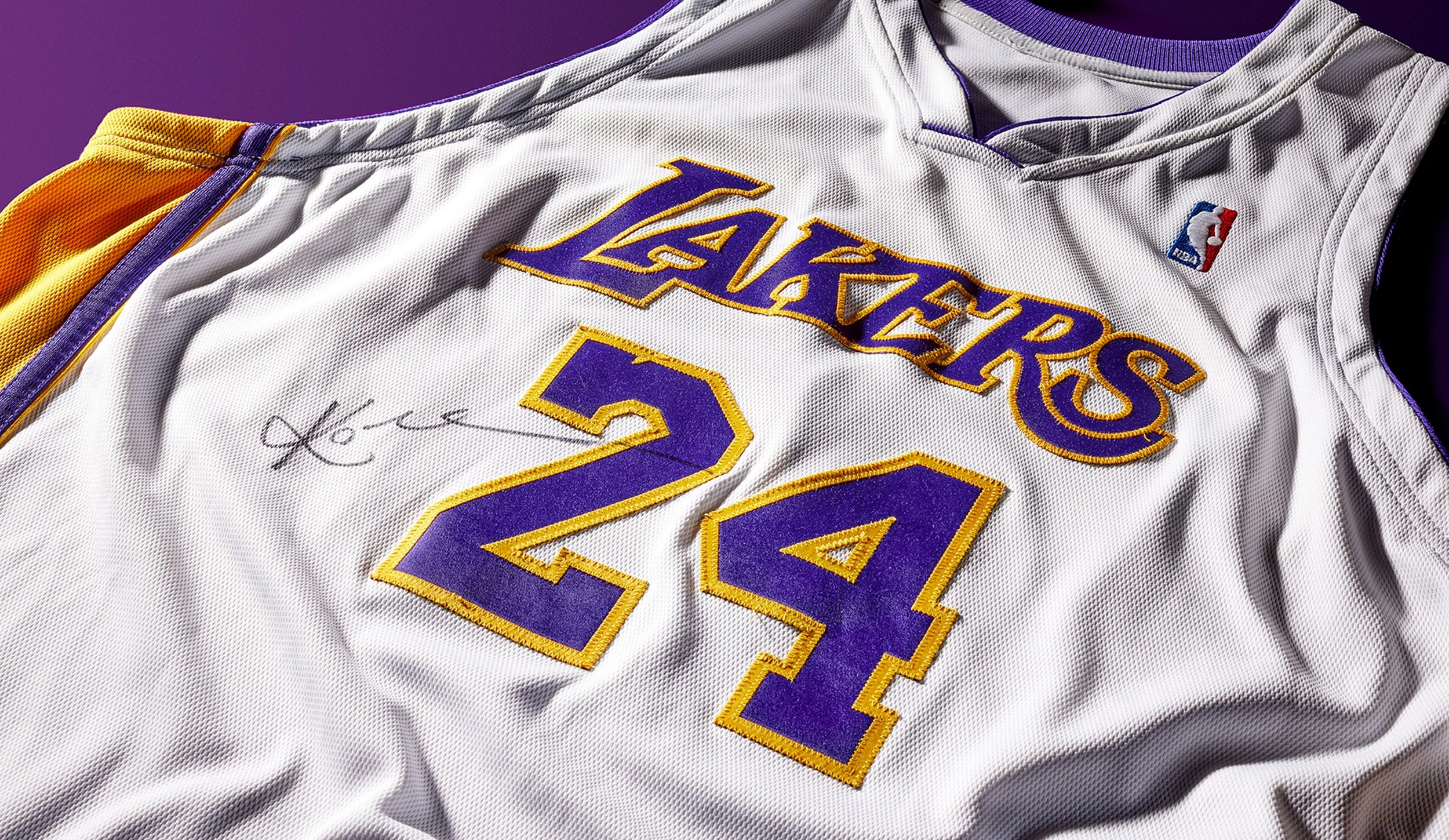 Close-up of a signed Los Angeles Lakers Kobe Bryant basketball jersey with purple and yellow accents, displaying the number 24 and an NBA logo.