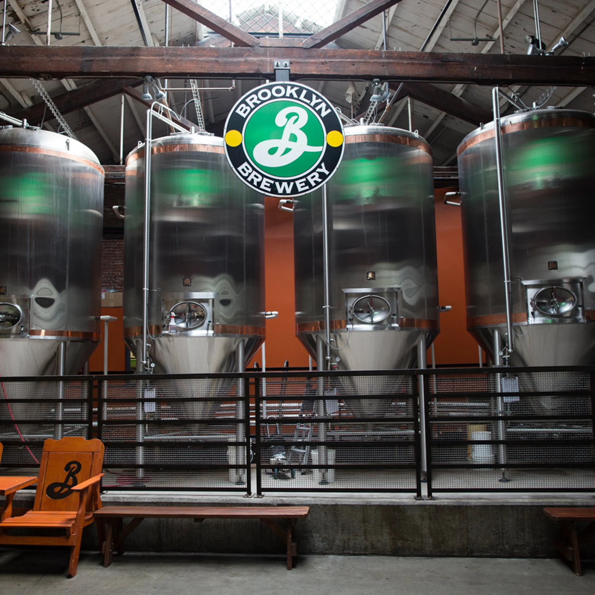 Interior view of a brewery with large stainless steel fermentation tanks, a wooden beam overhead, and a sign reading 'Brooklyn Brewery' hanging in front of the tanks.