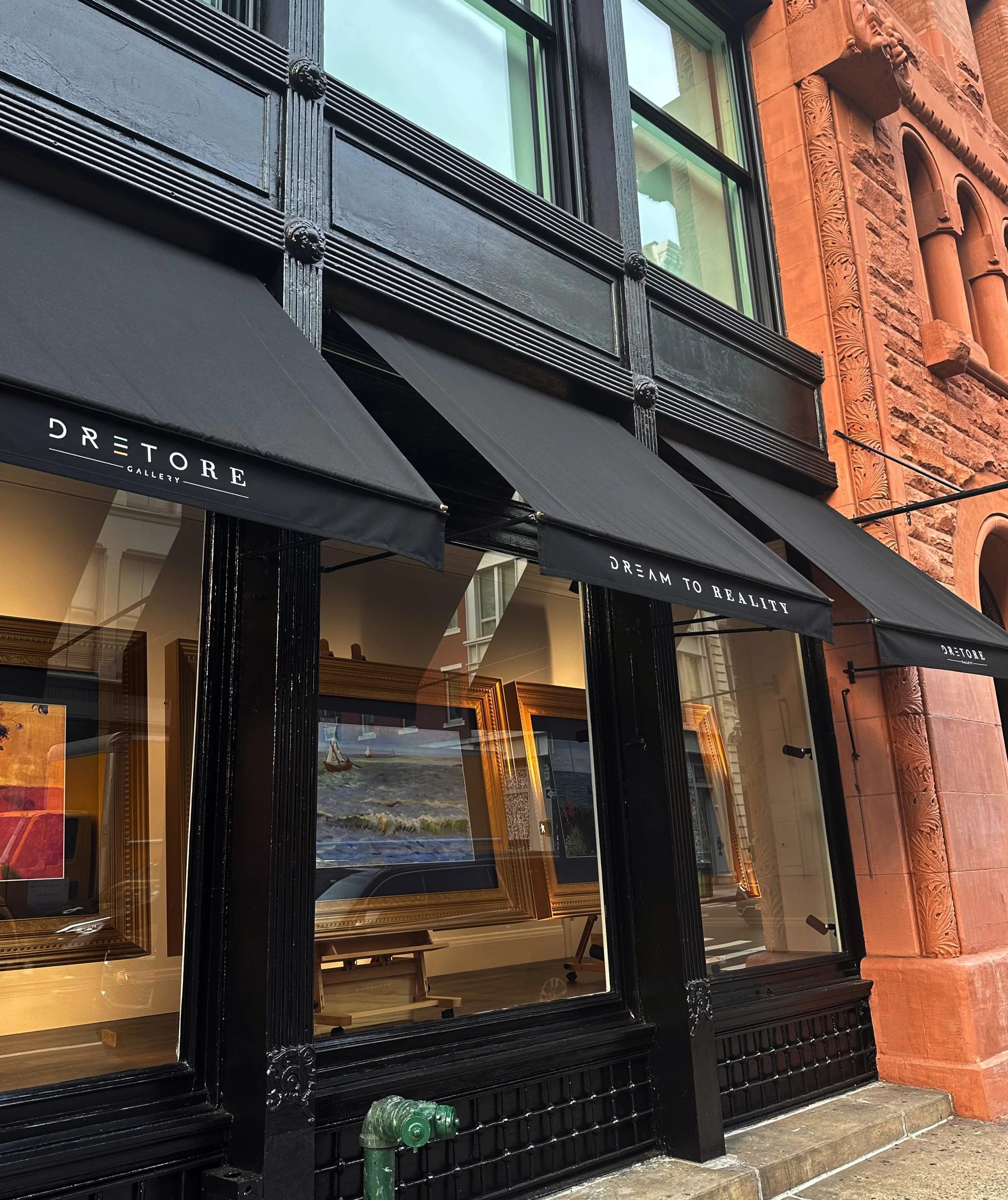 Exterior of a storefront with black awnings displaying the words "DREAM TO REALITY GALLERY." Large windows showcase framed artwork inside, including paintings and photographs. The building features ornate architectural details, including intricate carvings and a brick facade.