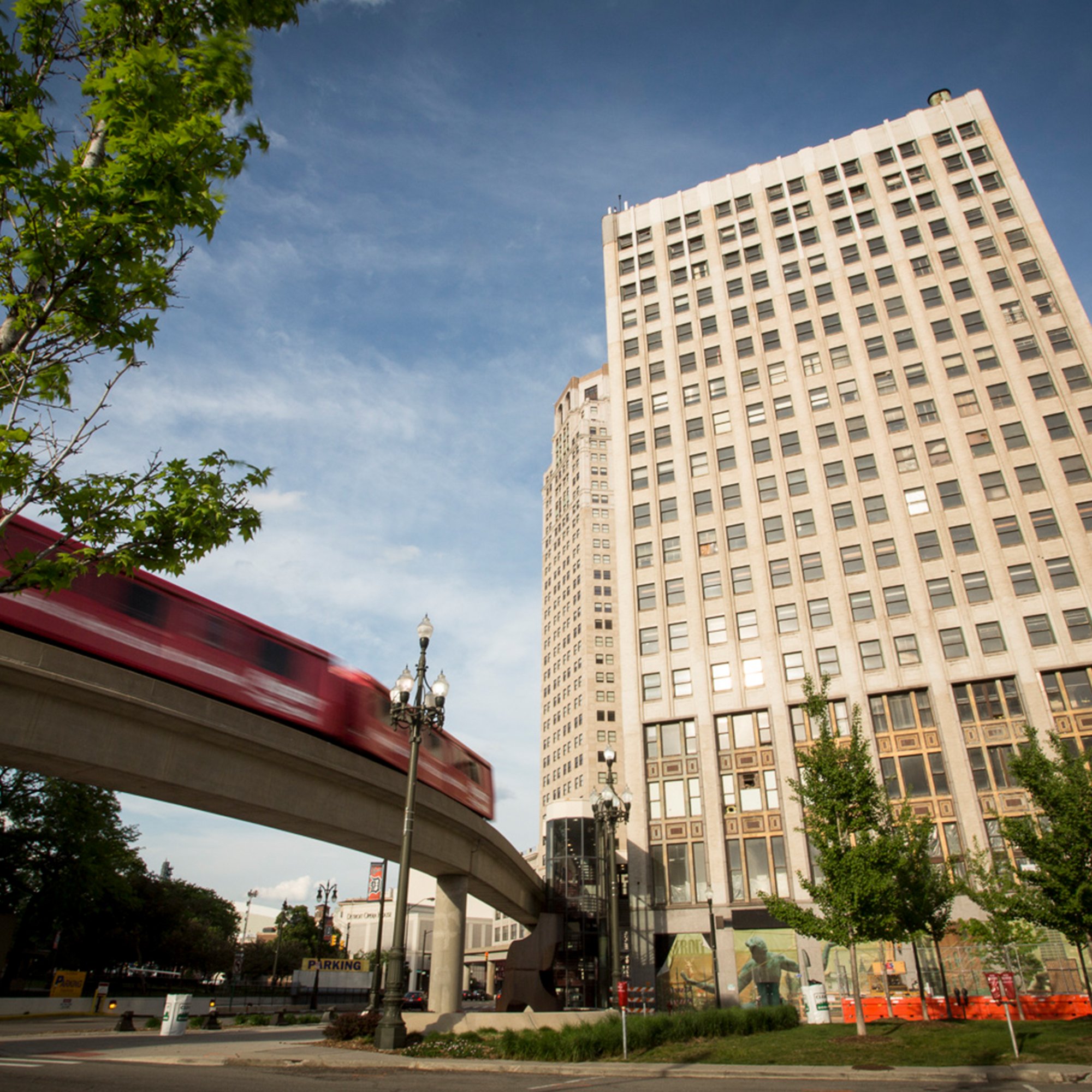 City street scene with a tall beige office building, green trees, streetlights, and a red transit bus in motion, under a partly cloudy sky.