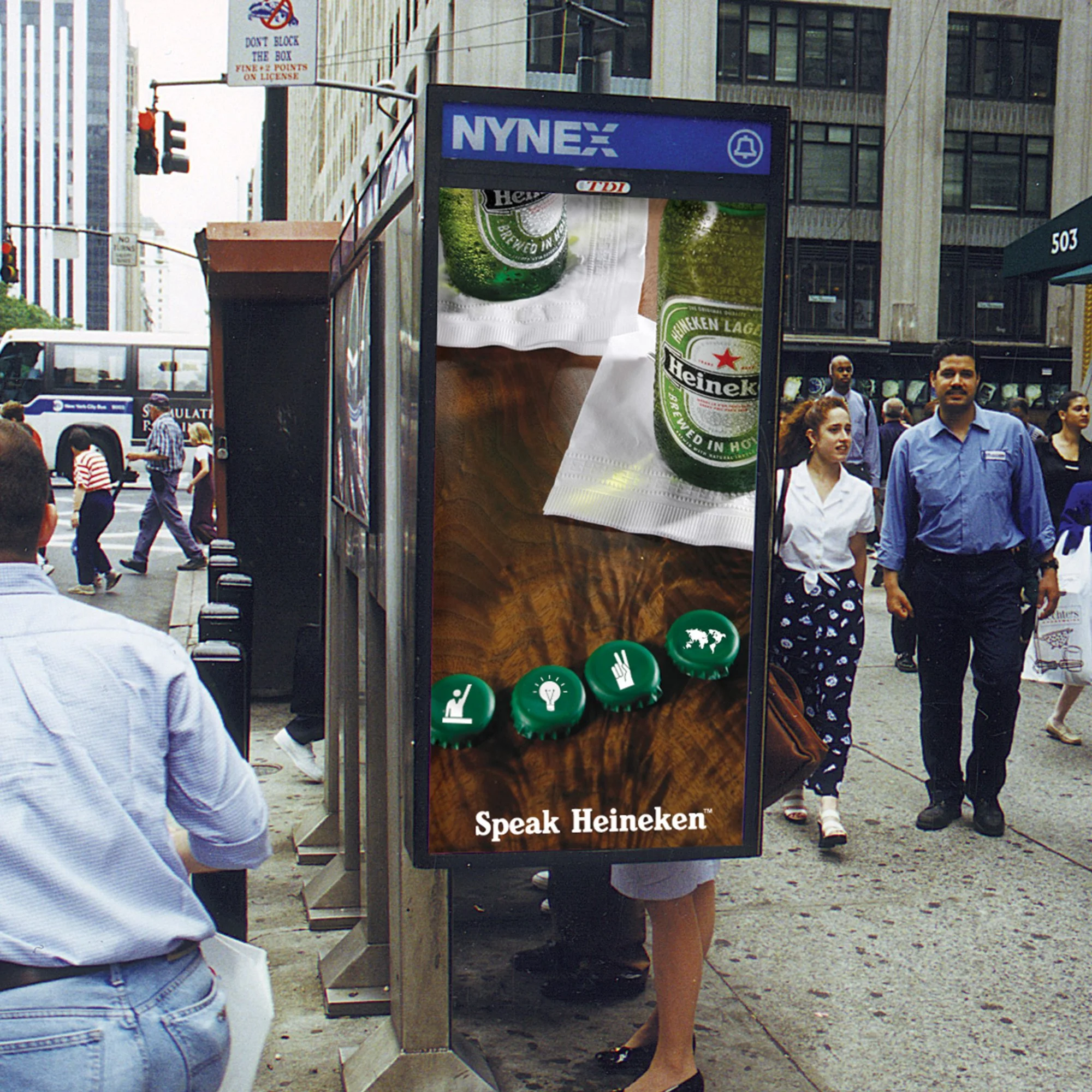 A city street scene with a digital billboard advertising Heineken beer, featuring an up-close image of a Heineken bottle and text that says 'Speak Heineken'. People are walking on the sidewalk, and buildings, traffic lights, and street signs are visible in the background.