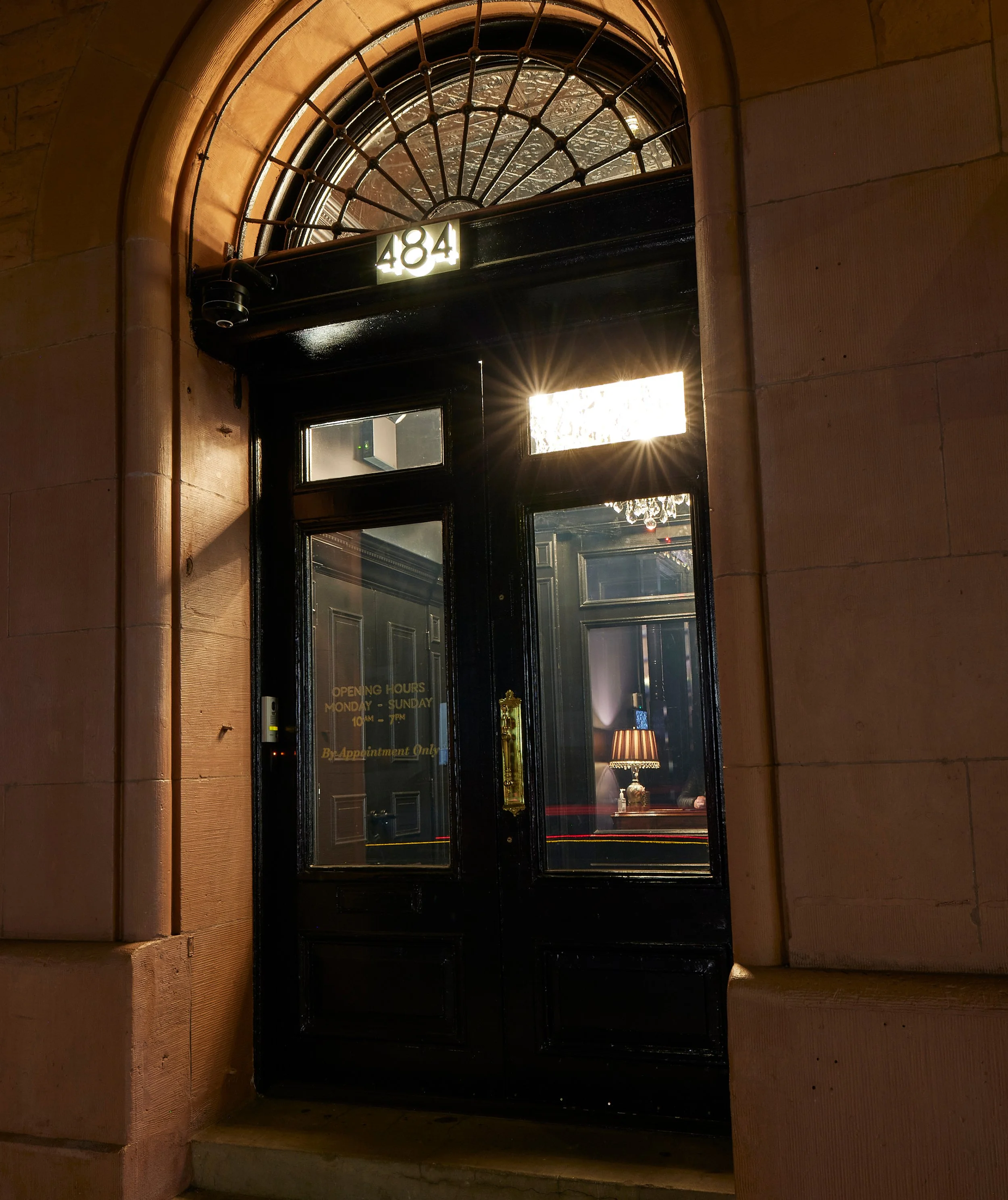 Hotel or building entrance with black door, glass panels, and a decorative semi-circular window above, displaying the address '484'. The door has a sign with operating hours and appointment-only notice, and inside is a chandelier and a wooden desk with a lamp.