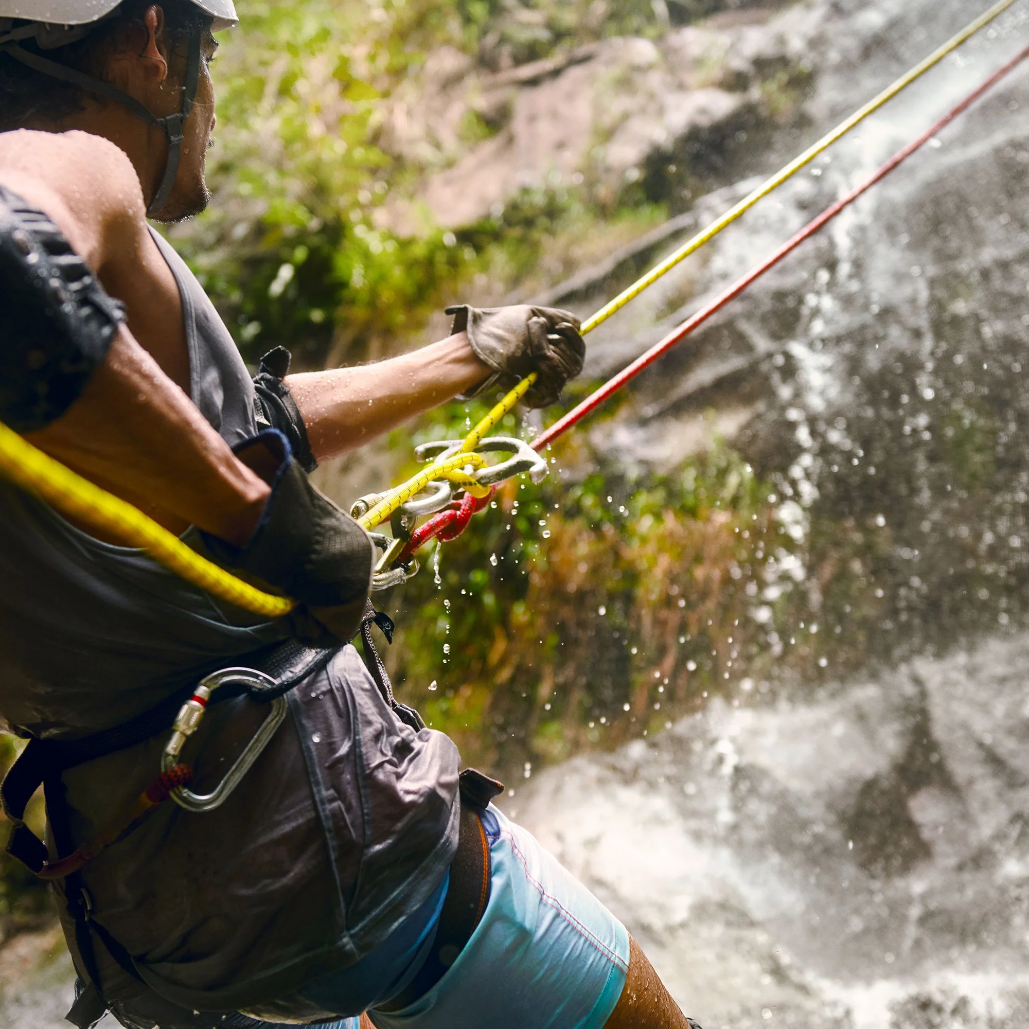 A person wearing a helmet and gloves rock climbing near a waterfall or wet rocks outdoors, with lush green foliage in the background.