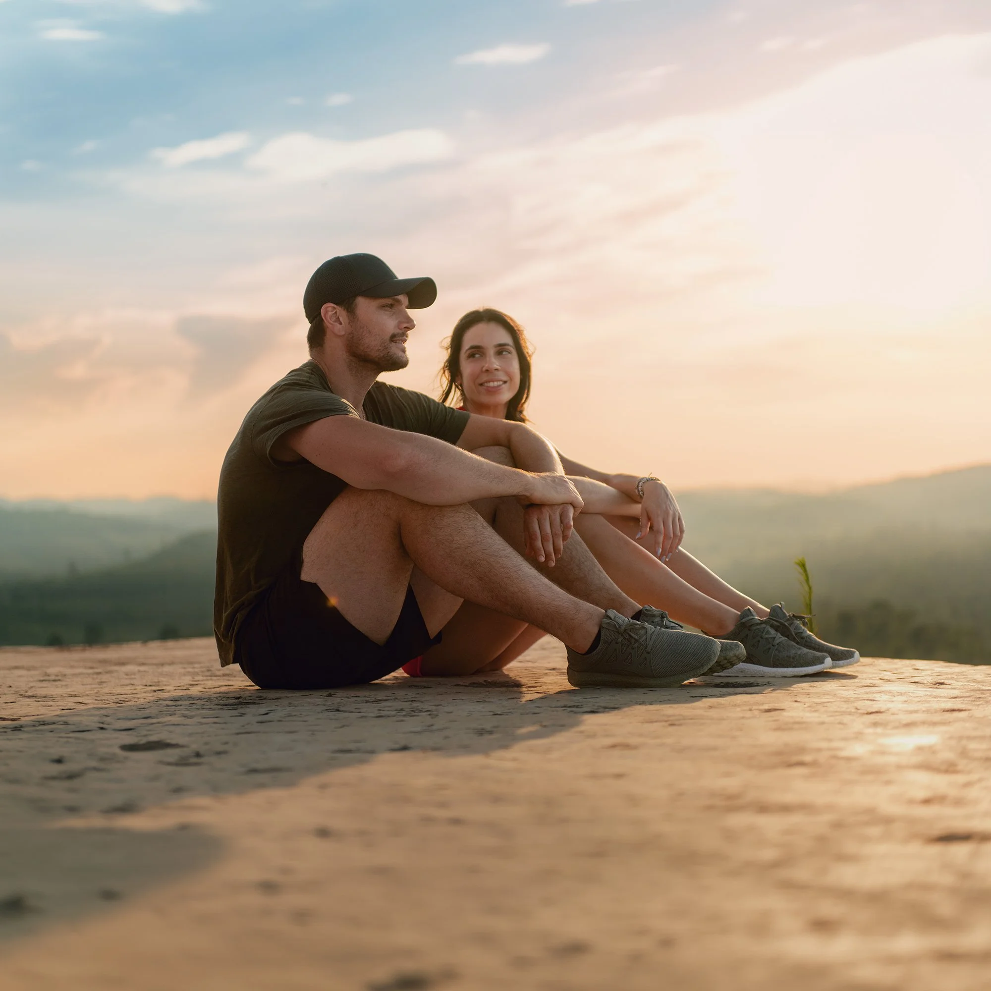 A young man and woman sit on a rocky surface at sunset, with the man looking to the left and the woman smiling while looking at him. Both are dressed casually and are outdoors in a natural setting.