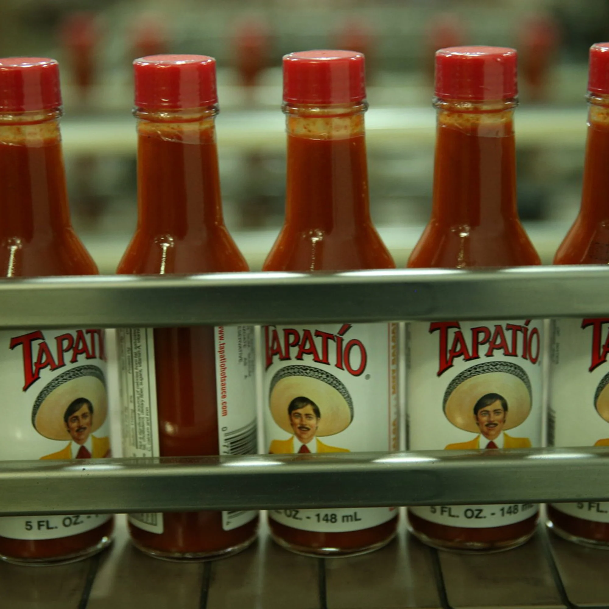 Row of small glass bottles containing a red sauce or condiment, with a female face and hair on the label, labeled 'Tapatío' hot sauce.