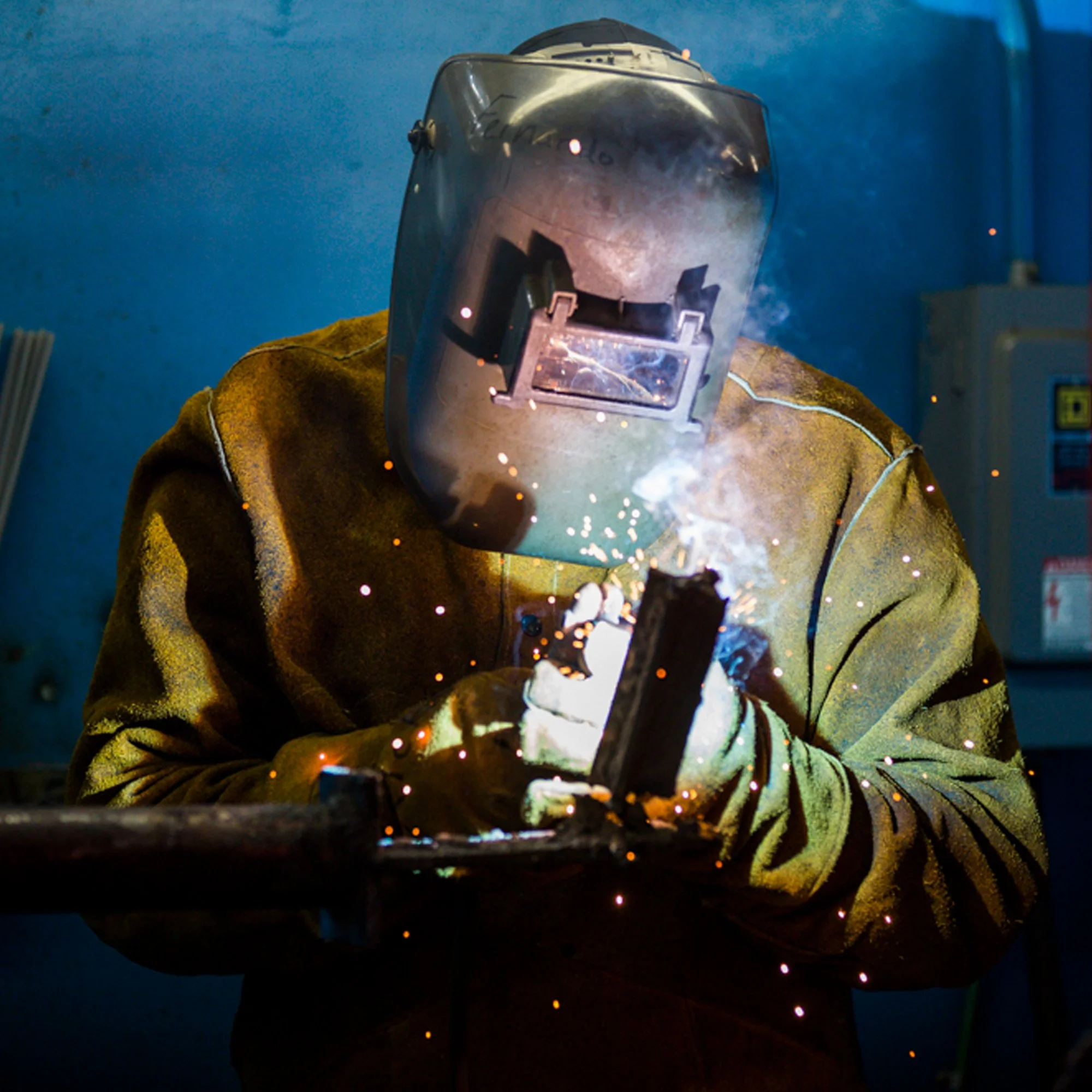 A person wearing protective welding gear, including a welding helmet and gloves, is welding metal, producing bright sparks.