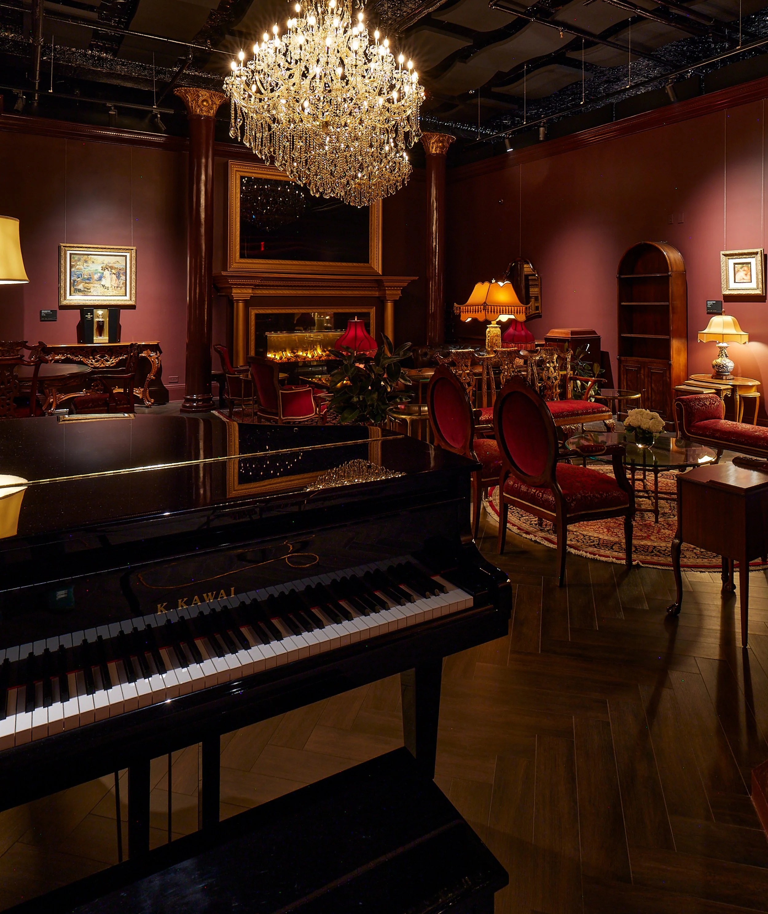 An elegant room with a large crystal chandelier hanging from the ceiling, a grand piano in the foreground, and vintage-style furniture including lamps, framed artwork, and a fireplace. The walls are painted a deep shade of red or burgundy.
