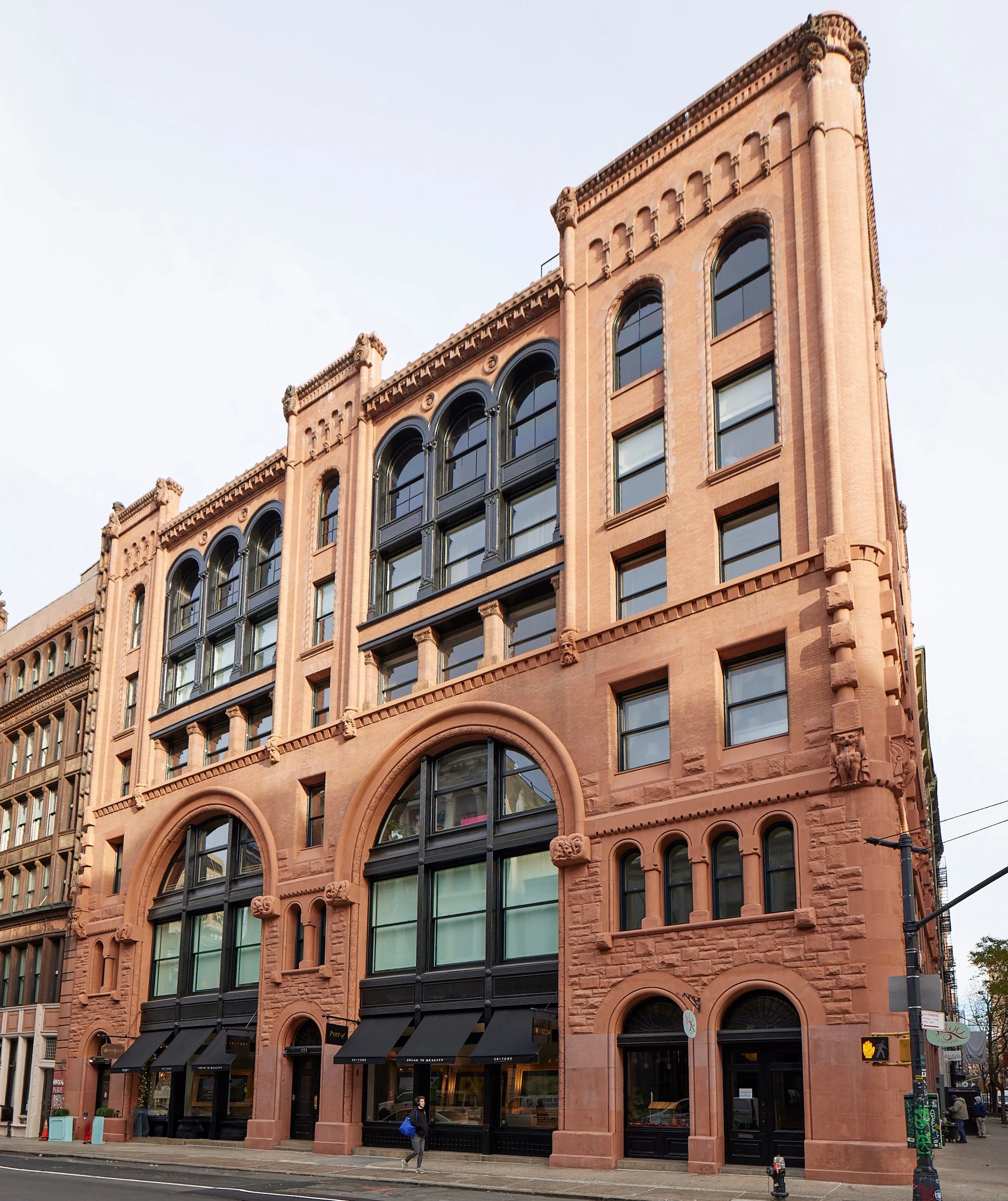 Historic pink sandstone building with arched windows and decorative architectural details, with storefronts on the ground floor and people walking on sidewalk in front.